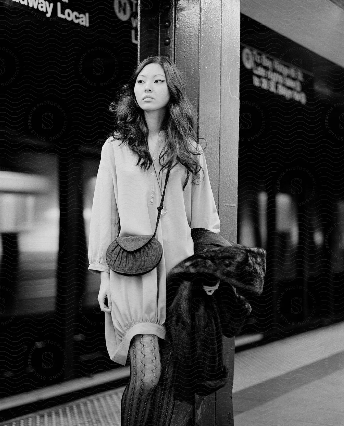 A young asian woman in a dress and stockings stands in a nyc subway station holding a coat