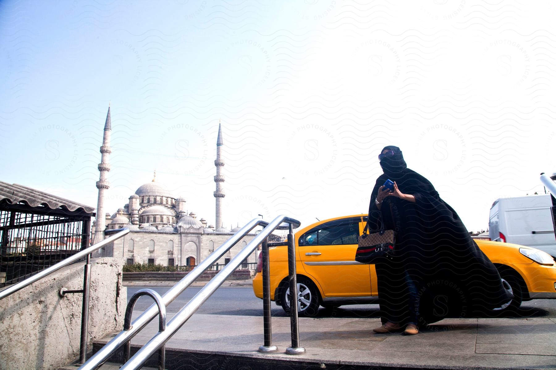 A woman in a black burqa walks out of a yellow car towards the stairs in front of a mosque