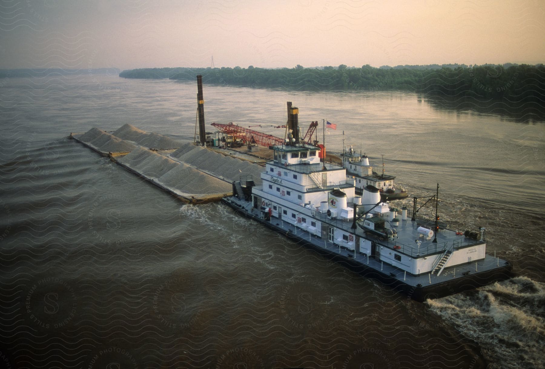 A river tugboat pushes a barge loaded with gravel down the river passing a wooded coastal bank