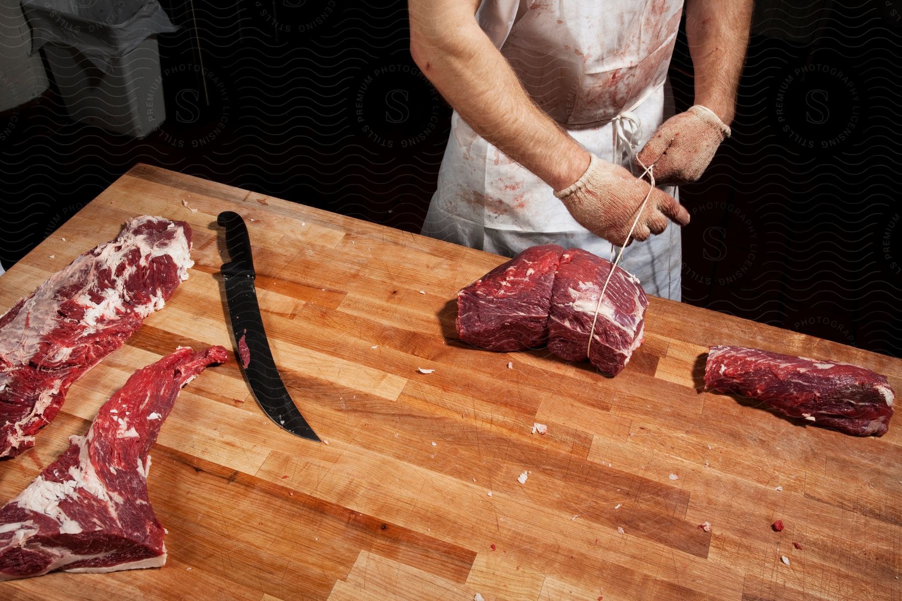 A man in gloves uses a knife to tie a slab of meat with a rope on a wooden table