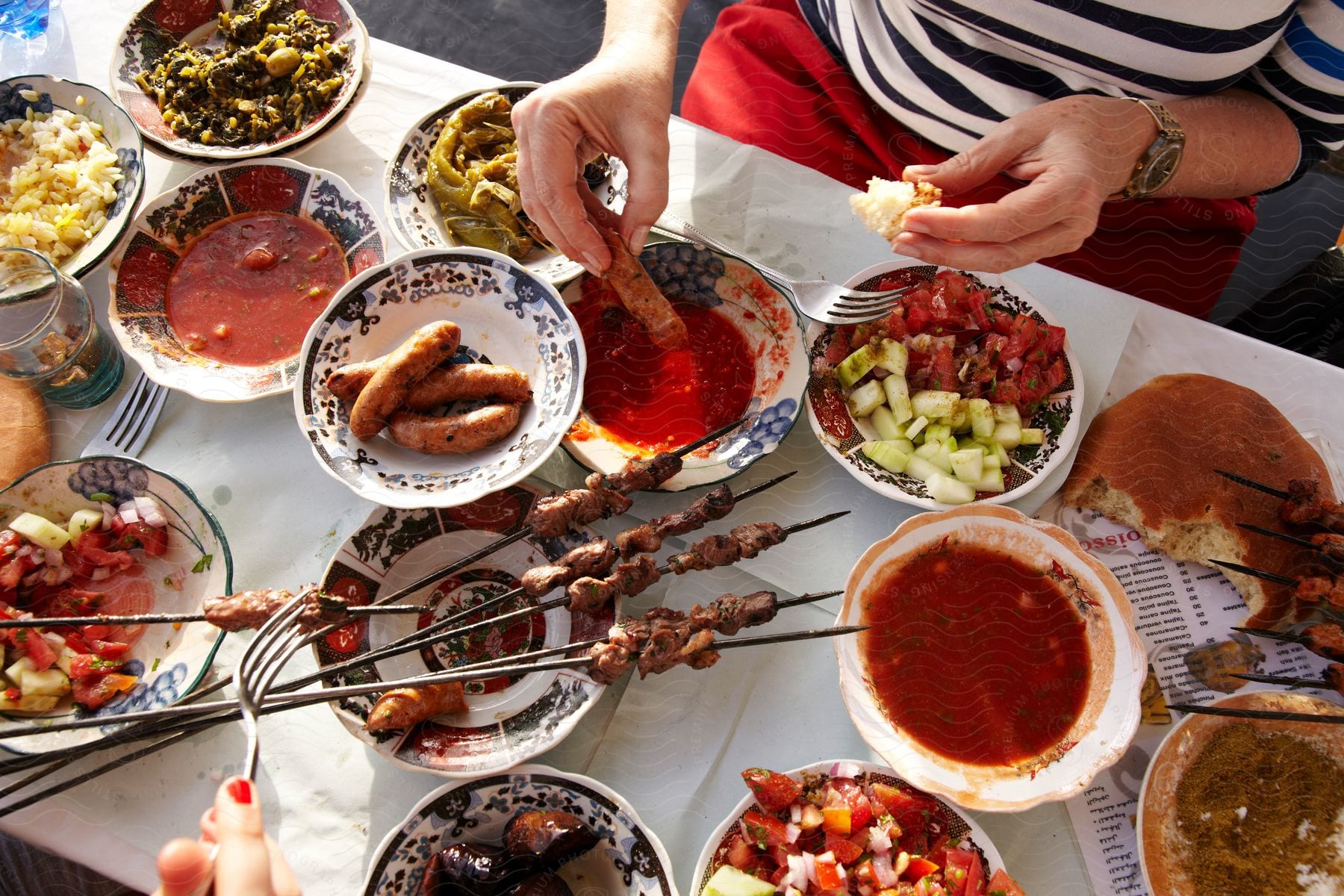 Top view of an outdoor table with bowls of sauces meat on skewers and a woman dipping a sausage into a bowl of red sauce