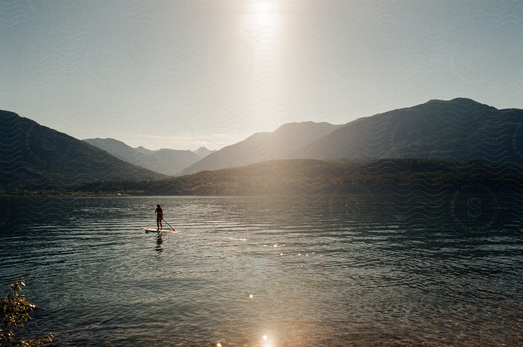 A person paddleboarding across a river under a hazy sky with mountains in the background