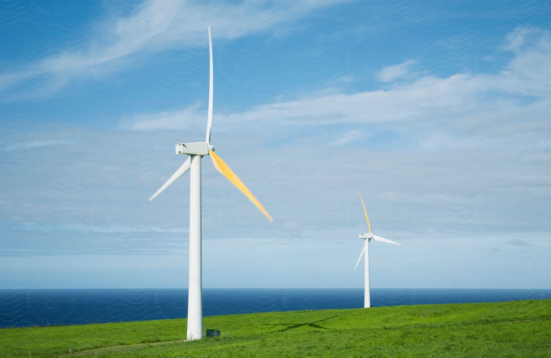 A group of wind turbines on a grassy hill near the ocean with a blue sky