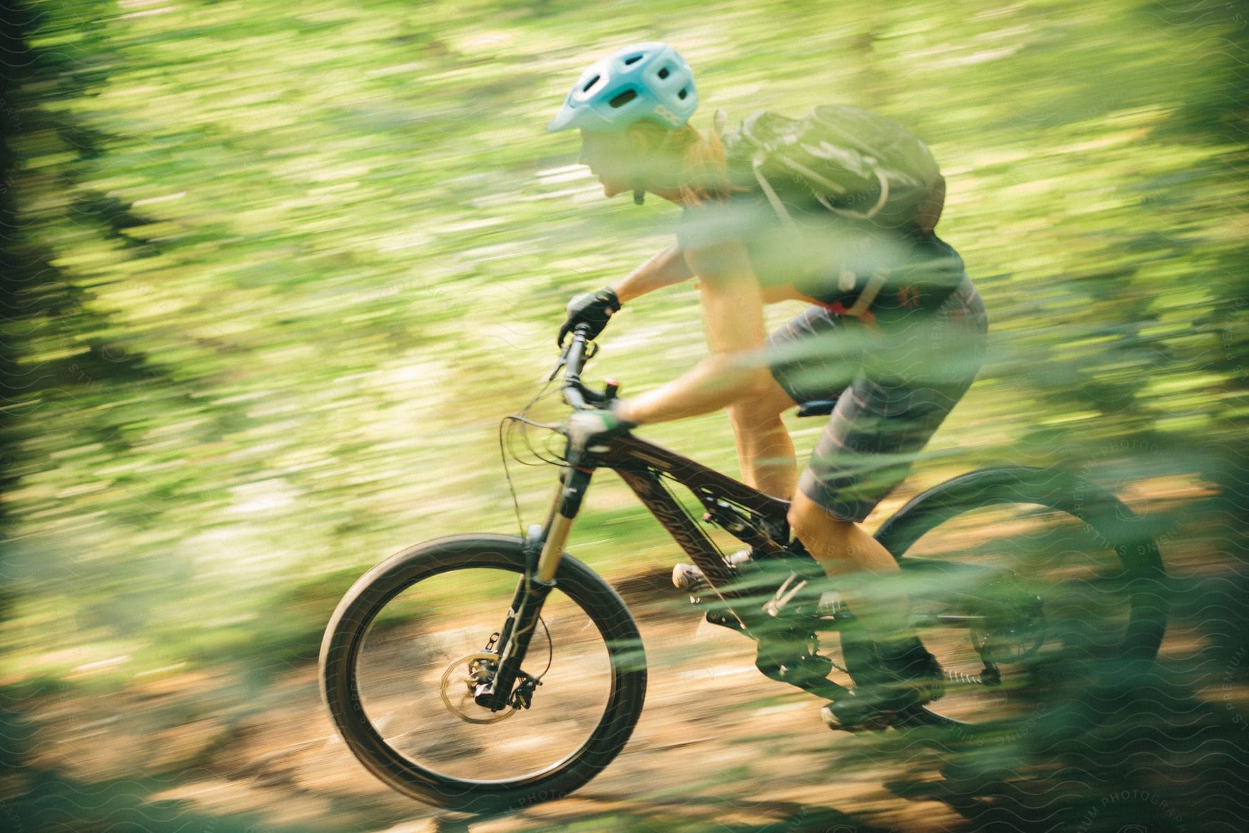 A blonde woman wearing a green tank top and grey shorts rides a mountain bike in a forest