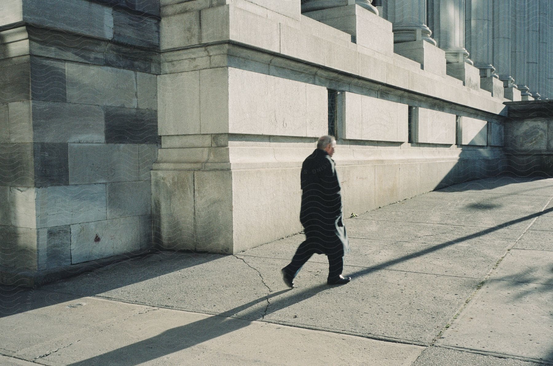 A man in a trench coat walking past a large stone building