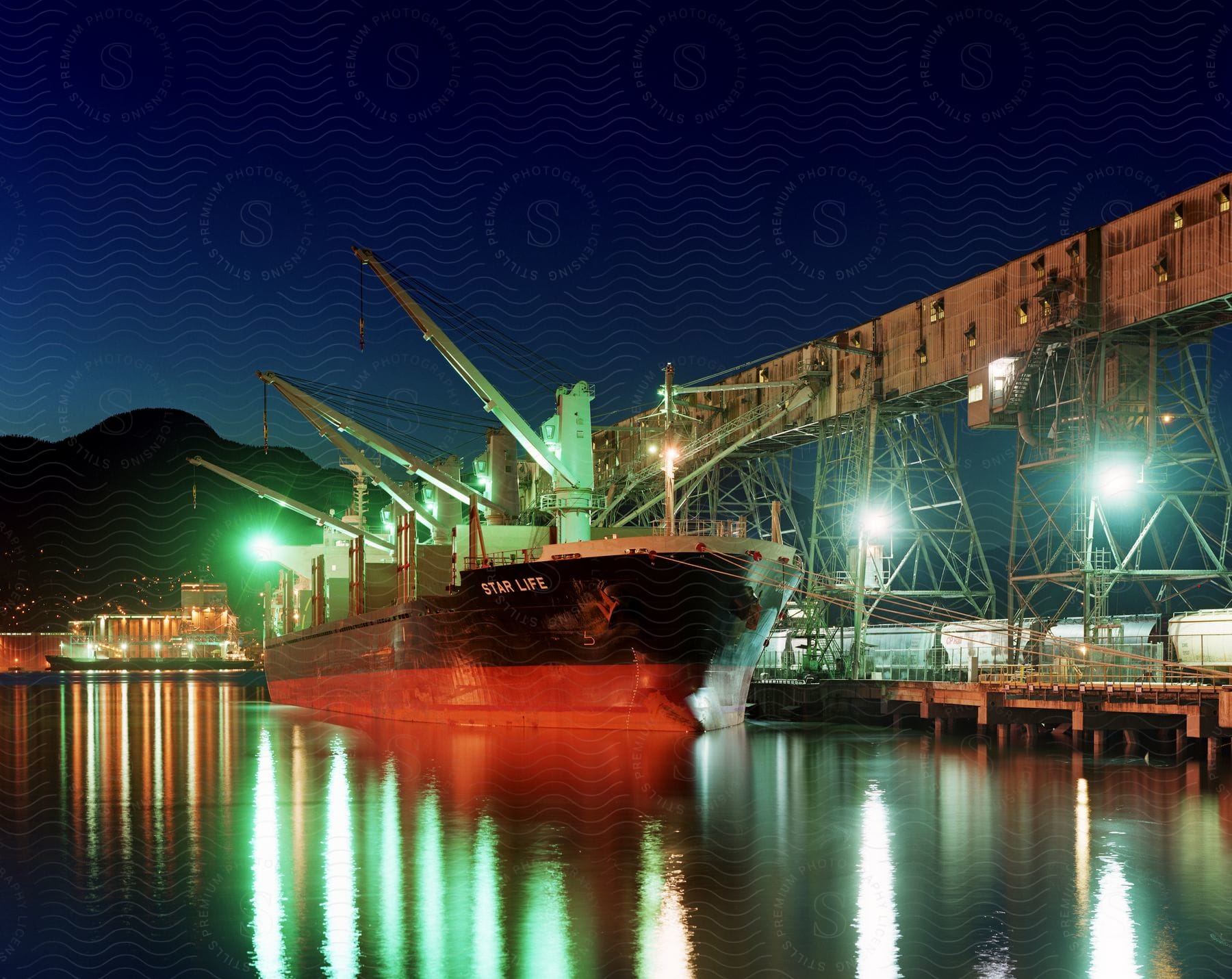 A cargo ship is docked in a brightly lit port at night