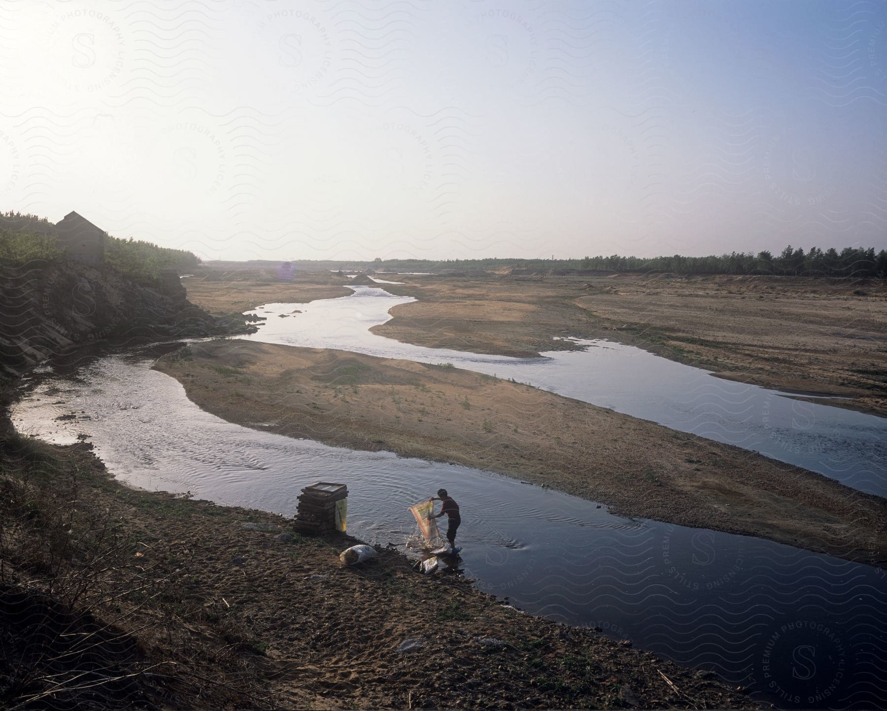 A man stands in a river fishing net in hand as the river flows away into the distance