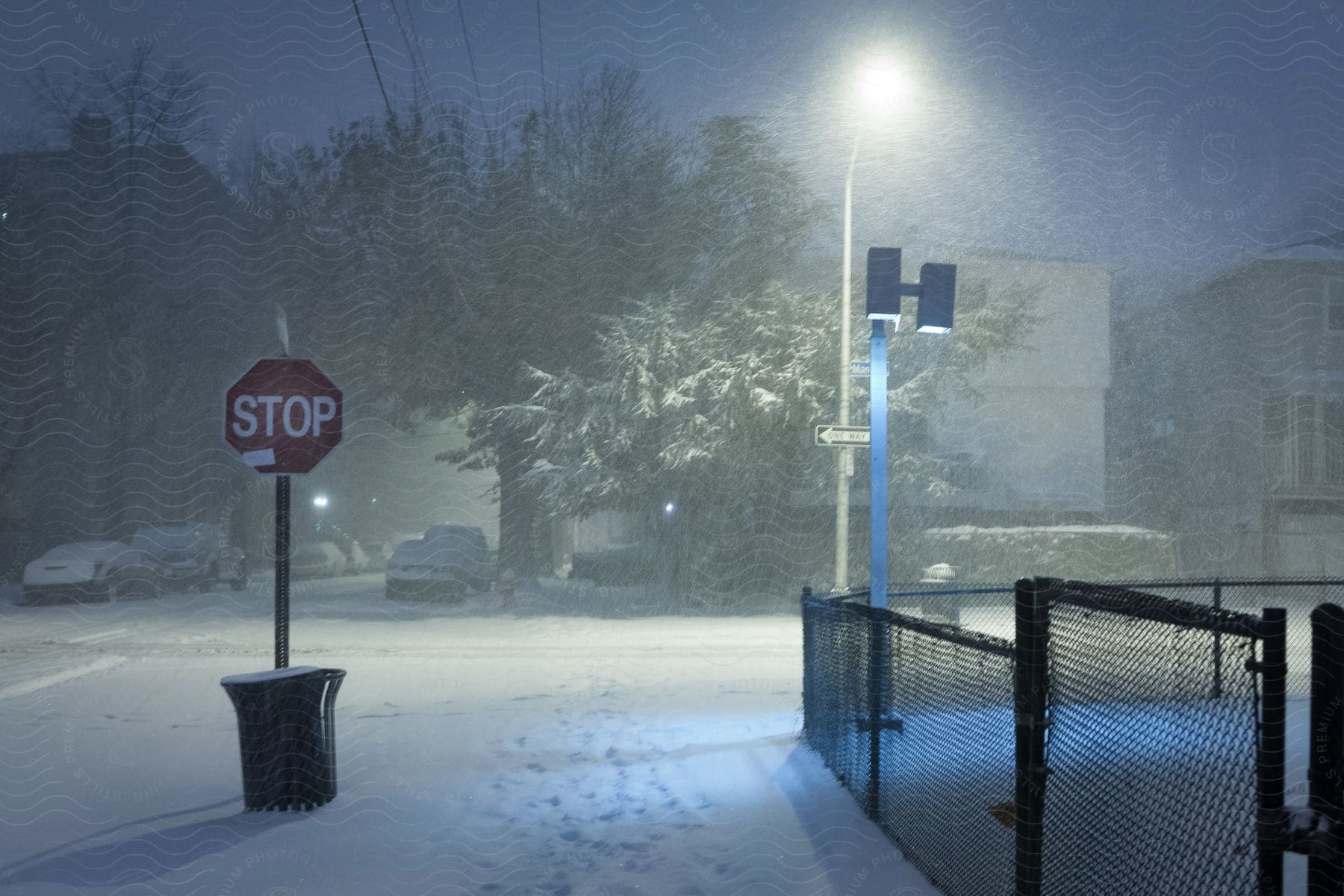 A snowcovered stop sign on a deserted street corner at night with a chainlink fence snowcovered vehicles trees and footprints illuminated by a street light in a blizzard