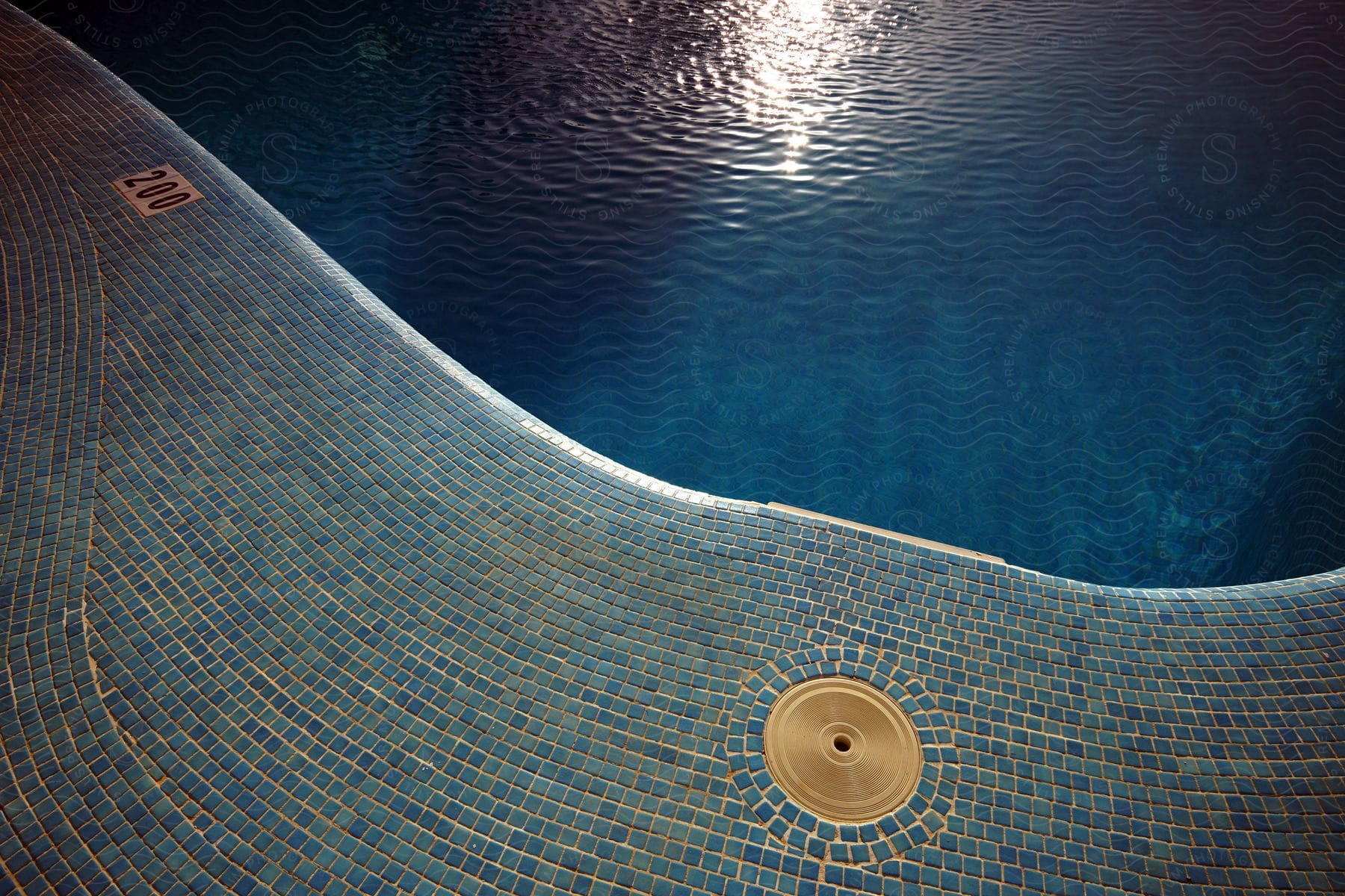 Blue mosaic tile at the edge of a pool full of dark blue clear water on a sunny day