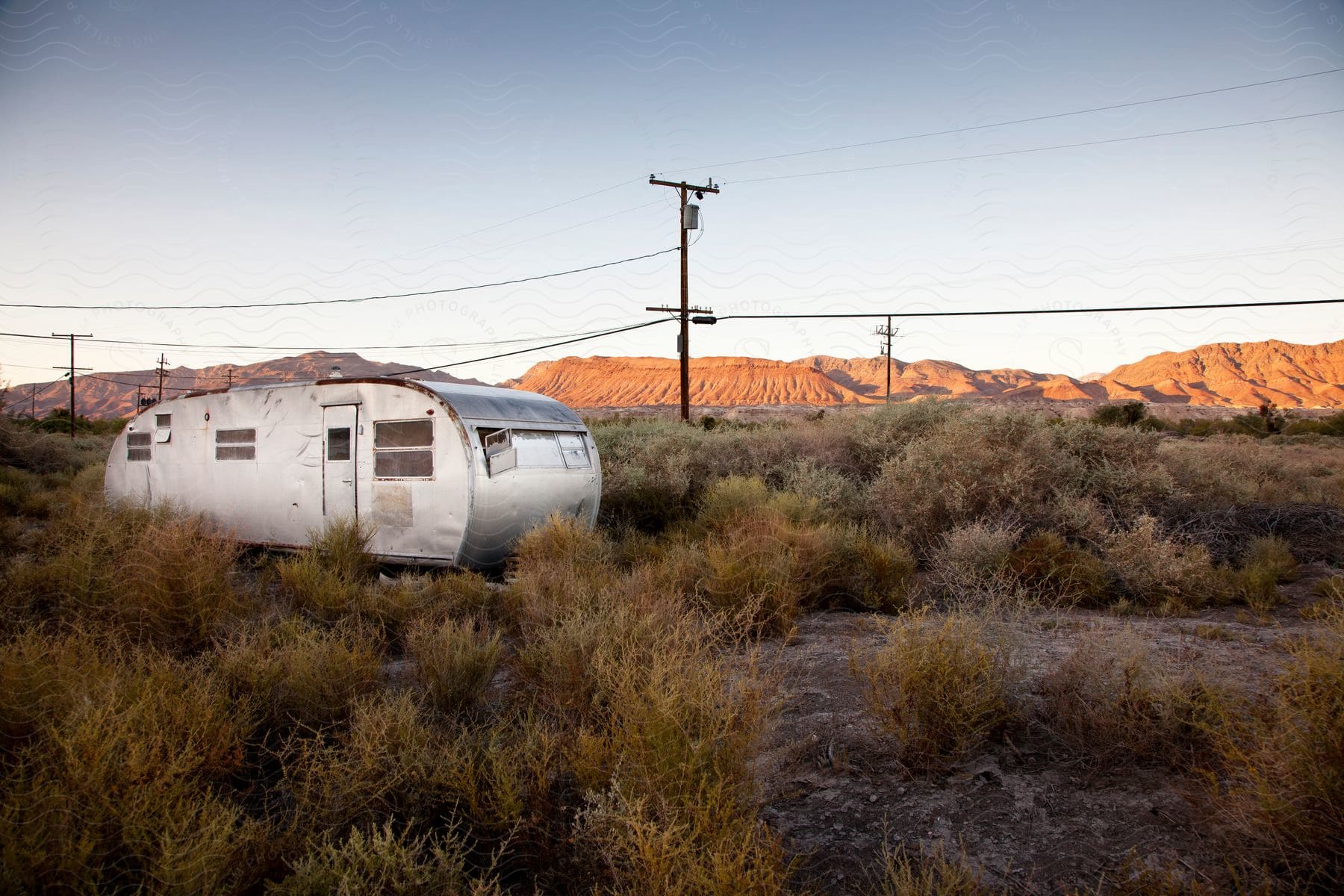 An old camper sits among plants with utility wires stretching across the landscape and mountains in the distance