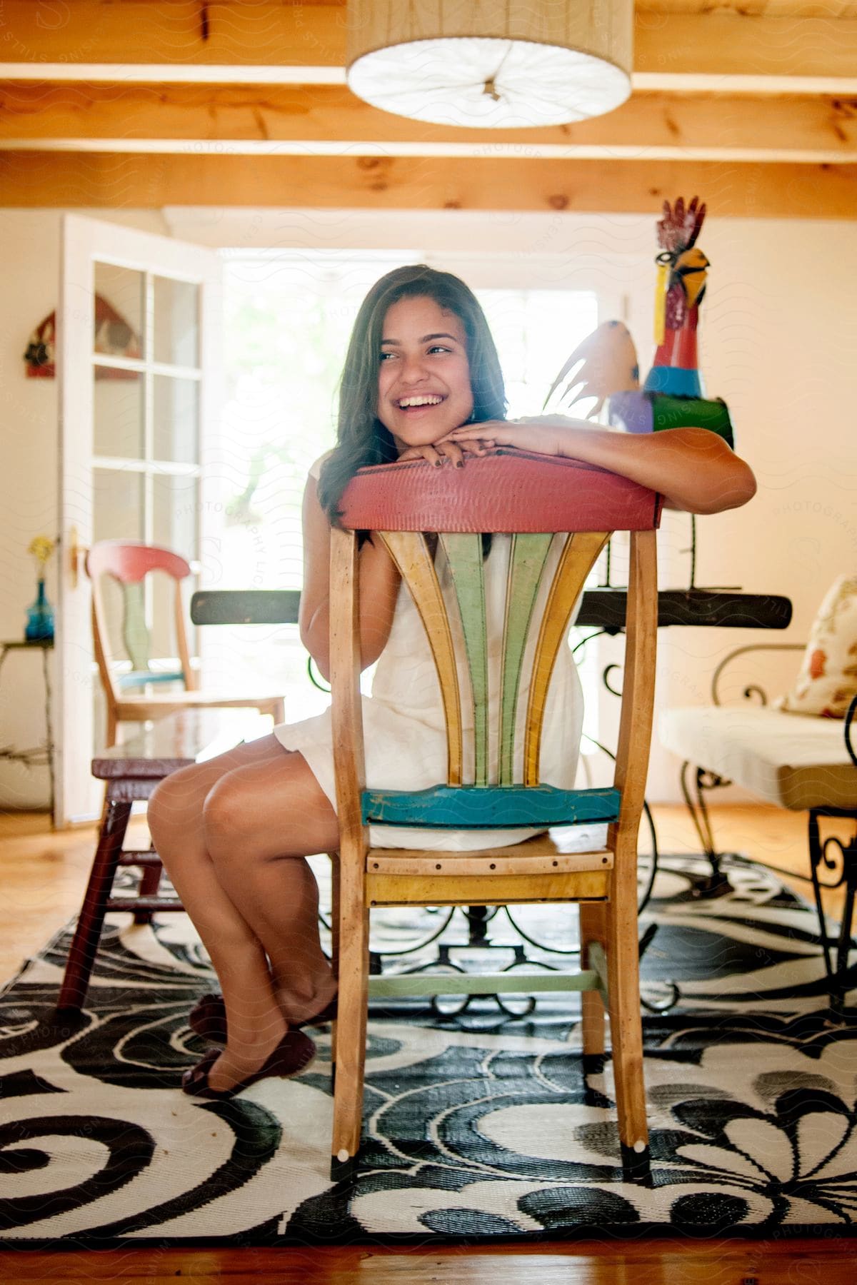A woman in a white dress sits in a colorful chair in a dining room smiling as sunlight comes through the open door