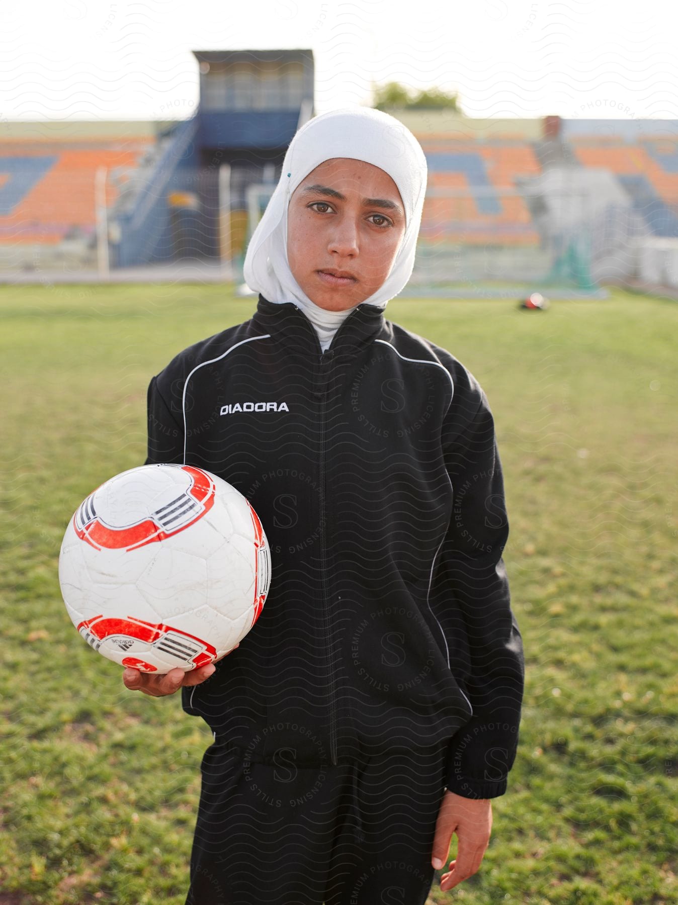 A woman wearing a headscarf holds a soccer ball on a soccer field