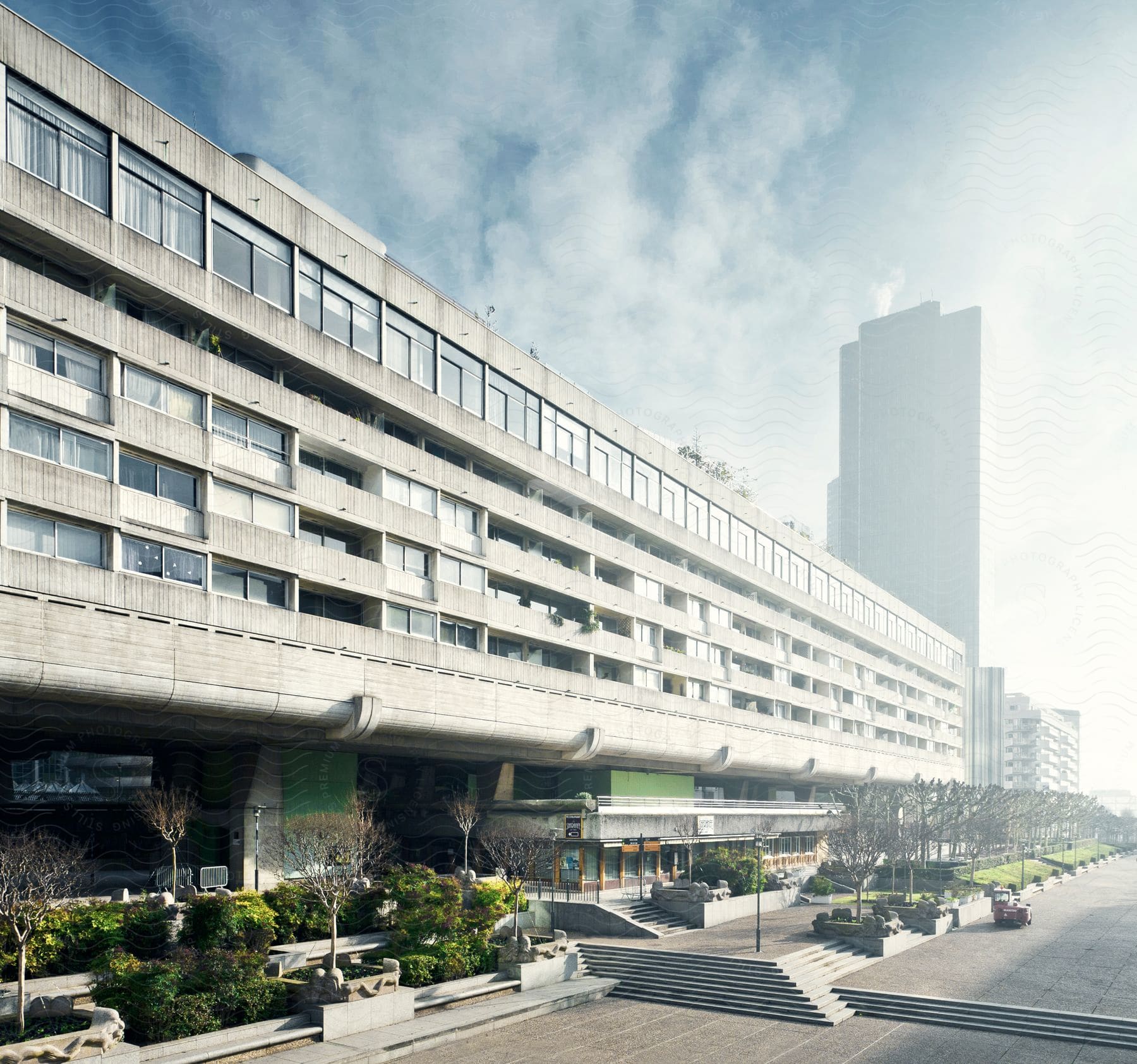 A large building with windows and a staircase stands among trees a distant skyscraper and clouds