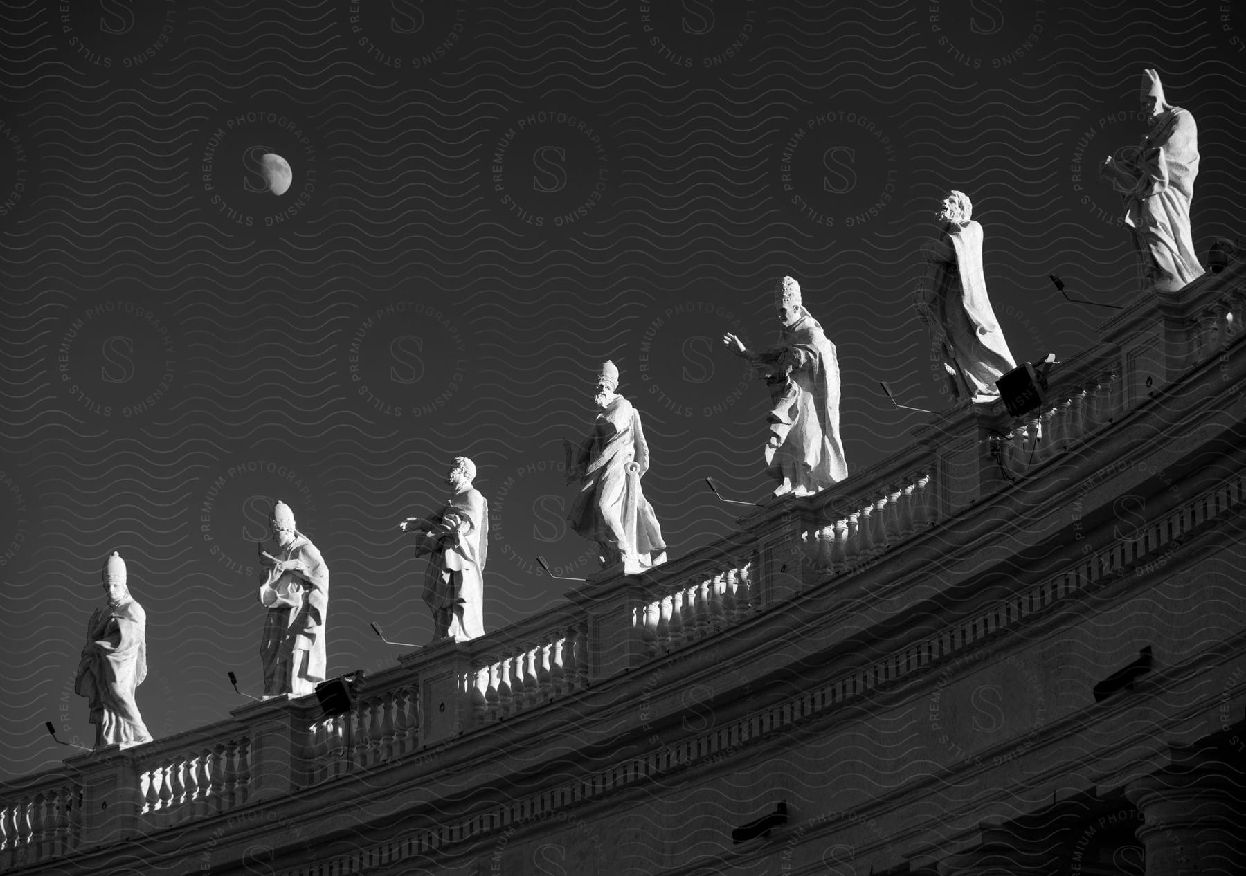 A row of statues on the balcony of a classical style building with a sky and moon in the background