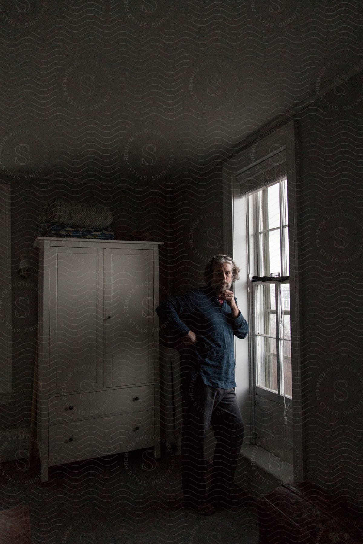 A man in his 50s standing in a dark bedroom next to a window with light coming in