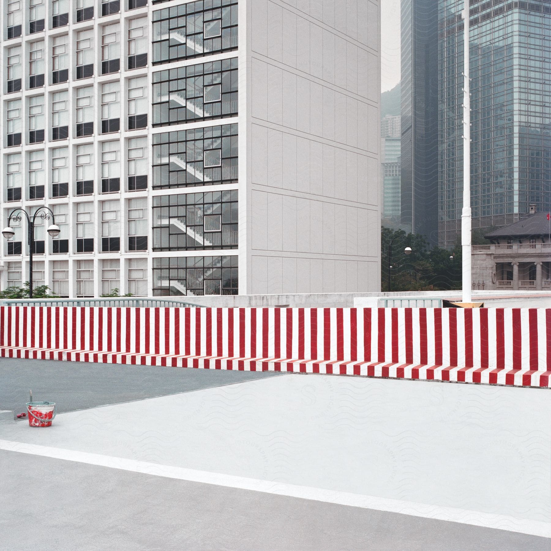 Sidewalk with red paint bucket and striped cement barrier in front of modern building with reflective windows