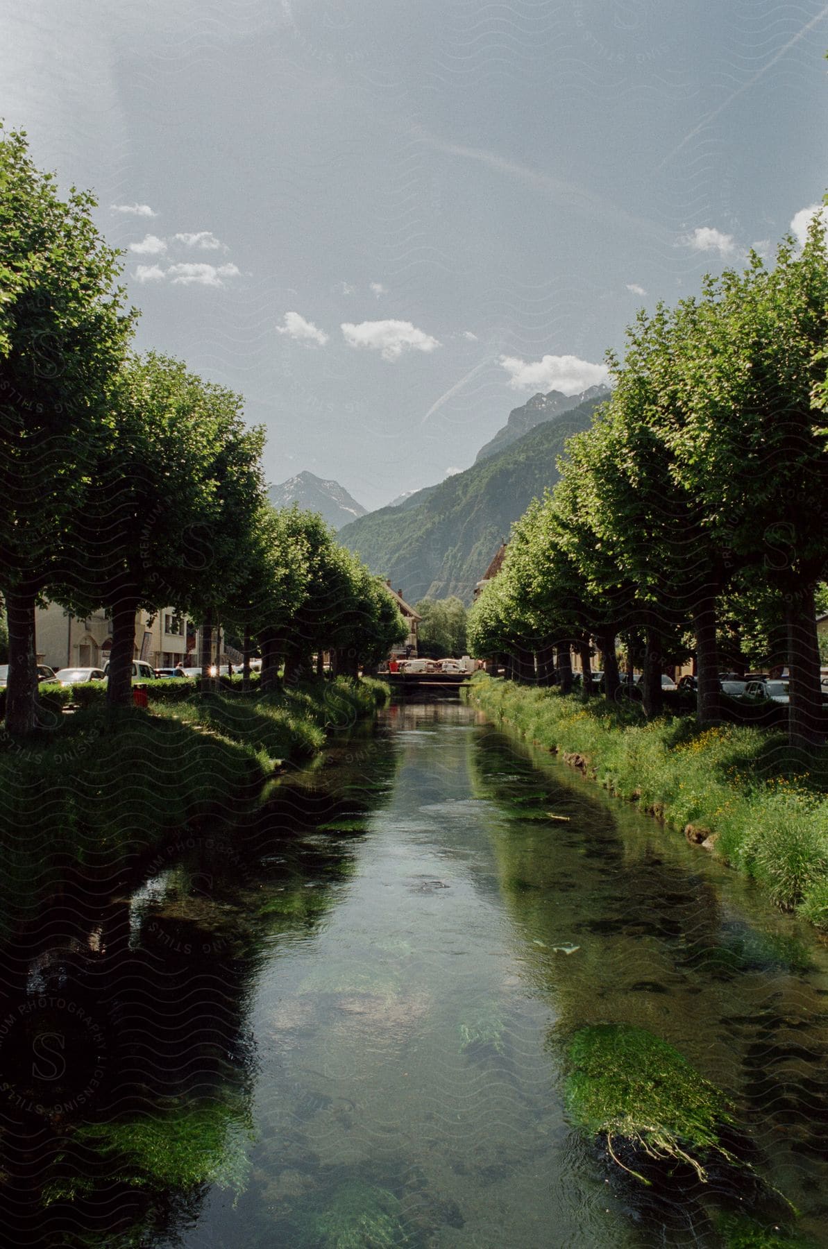 A River Flowing Through A Path Surrounded By Green Trees Buildings And A Distant Mountain