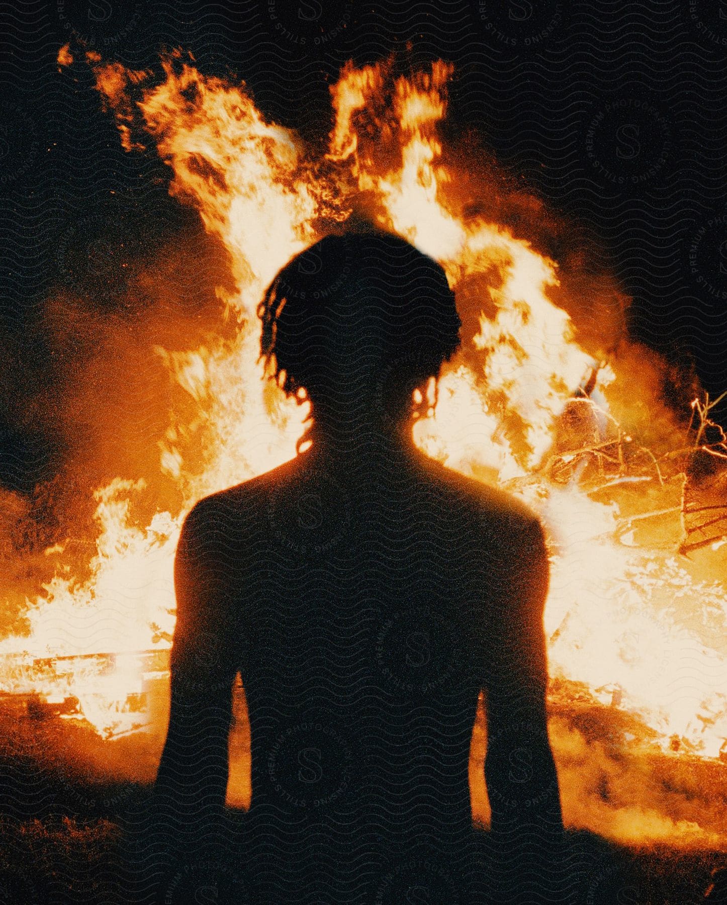 A man looks at a bonfire at night with his silhouette against the flames