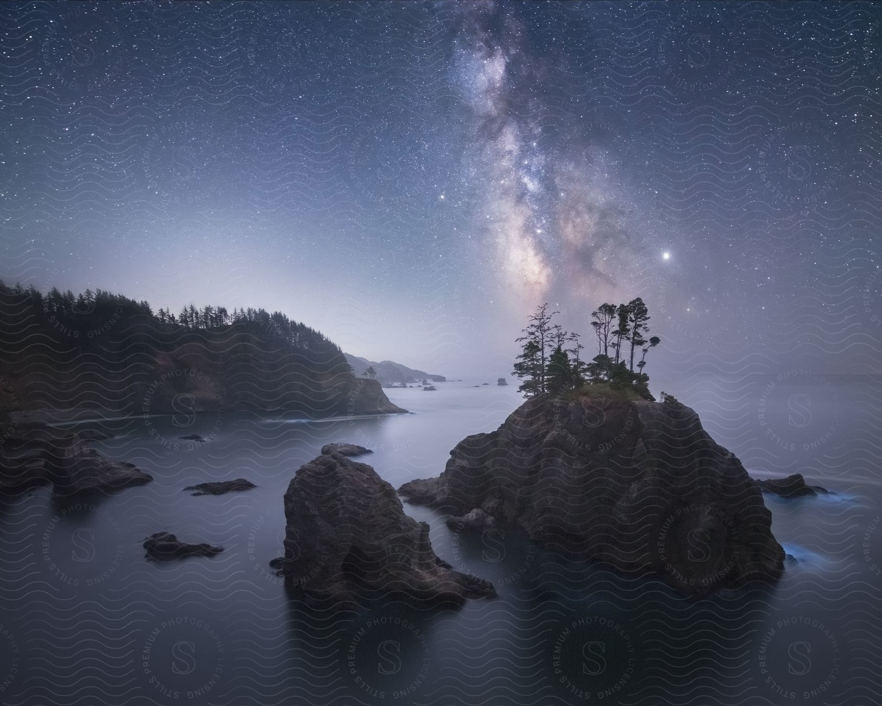 A calm coastal scene with rocky cliffs trees and a galaxy in the night sky