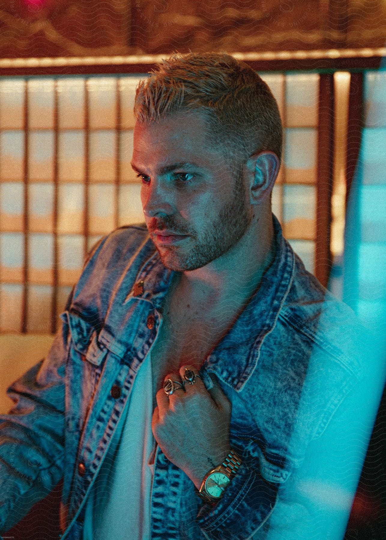 A man with blonde hair and a beard wearing a white vneck tshirt denim jacket wrist watch and rings on his left hand