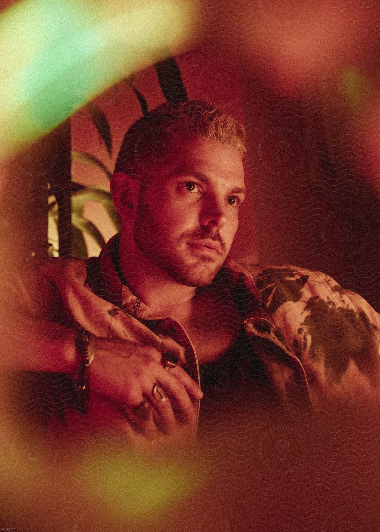 A blond man in a jacket posing with his head turned to the right is bathed in orangetoned lighting