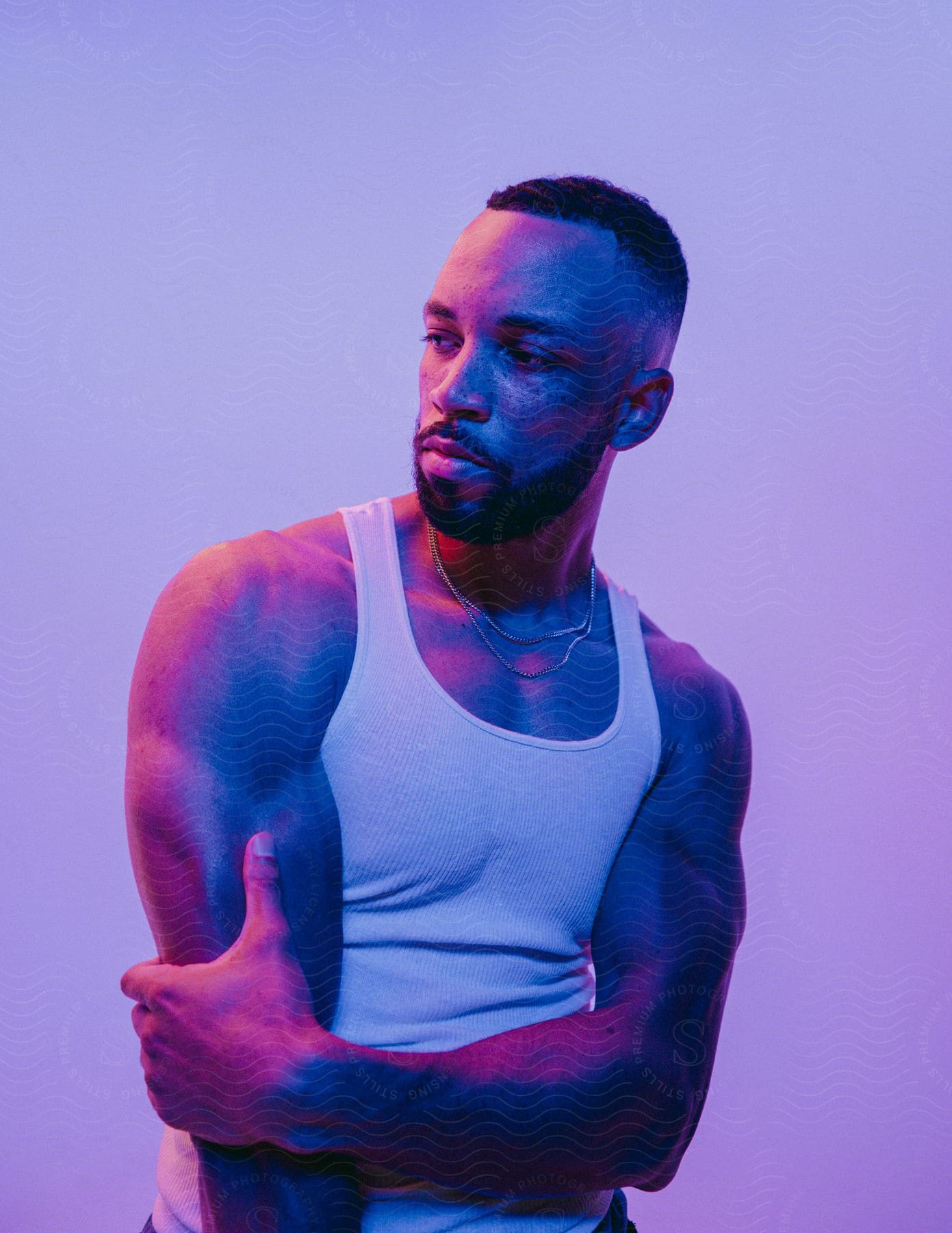 A black man with a beard and a white tank top is posing with crossed arms while purple and red light shines on him