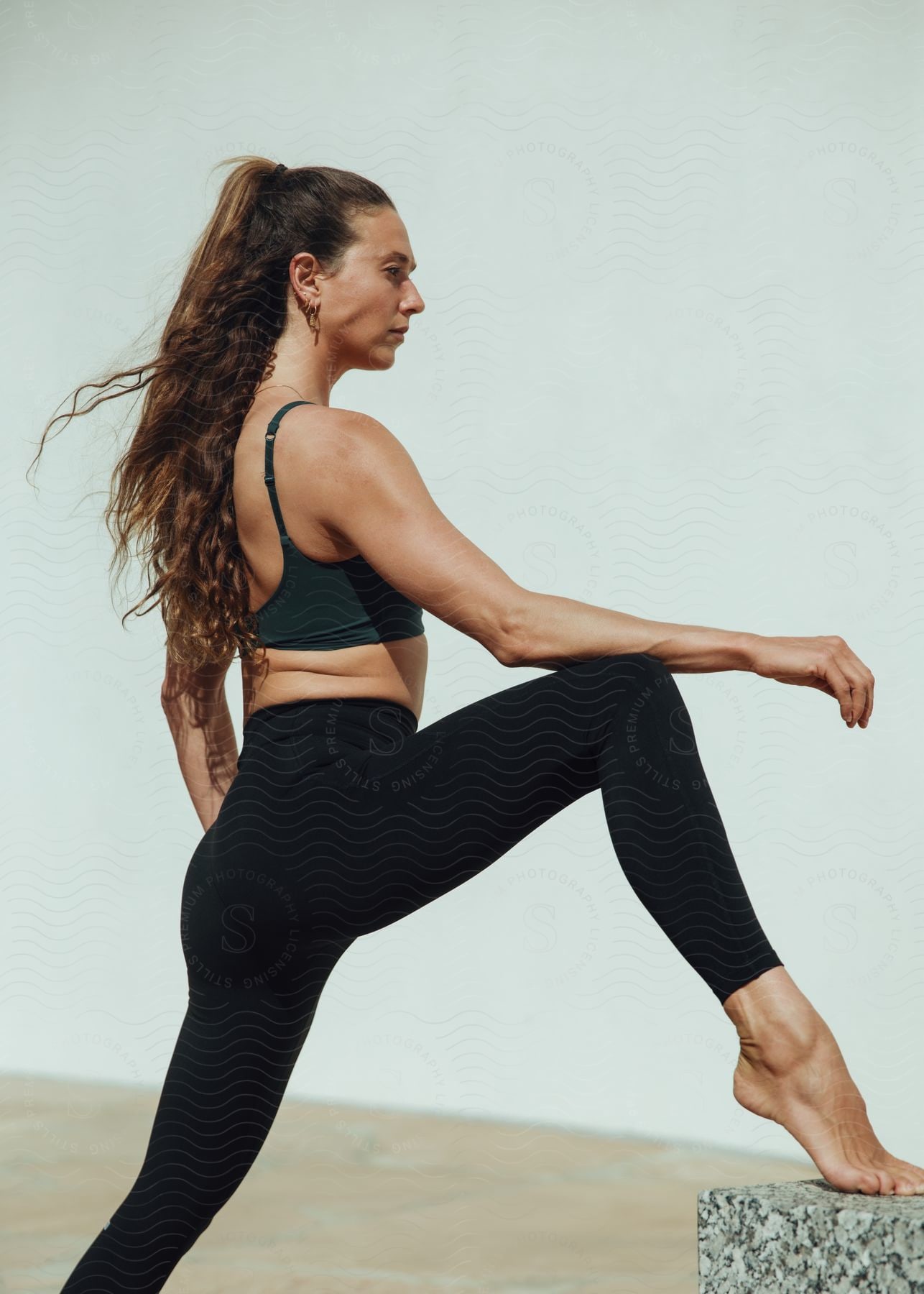 An older woman in black sports bra and yoga tights is balancing on one foot on a stone block