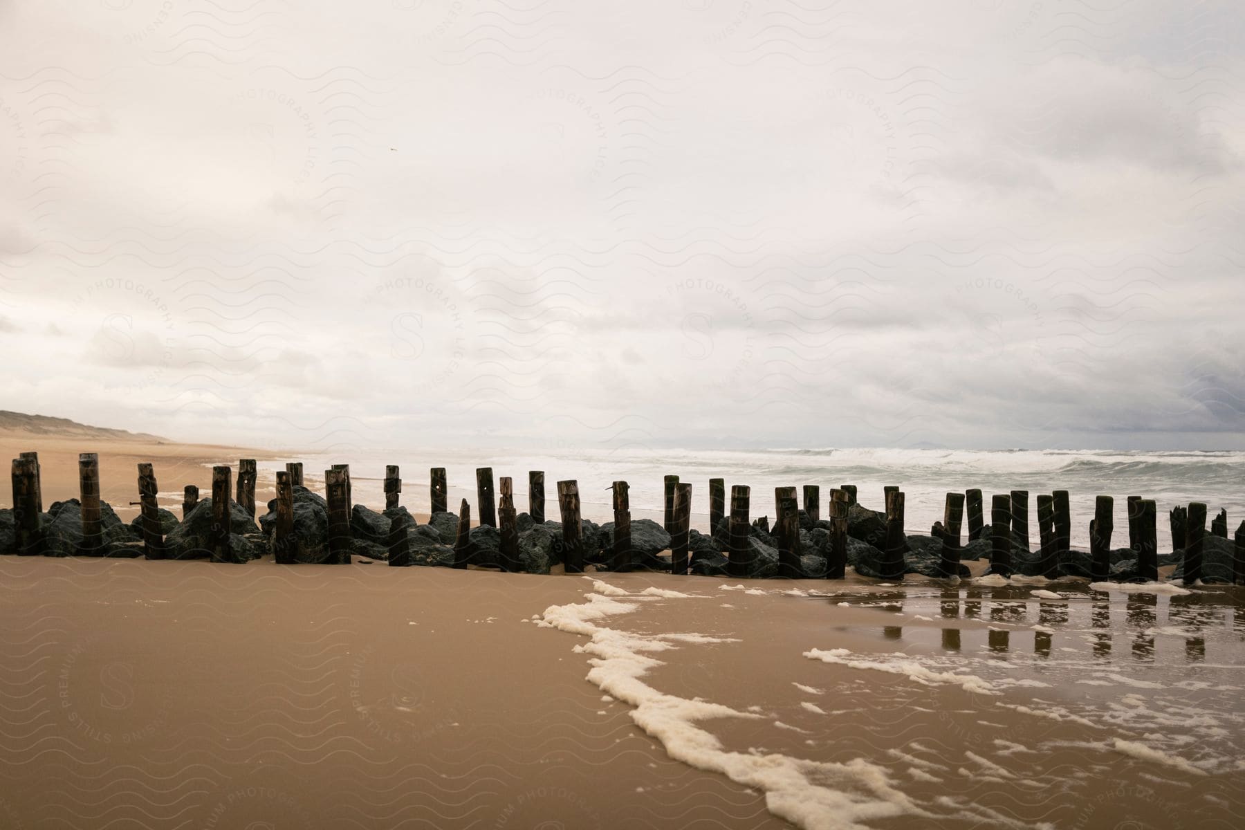 Piling and boulders are stacked in a line on the ocean shore under cloudy skies