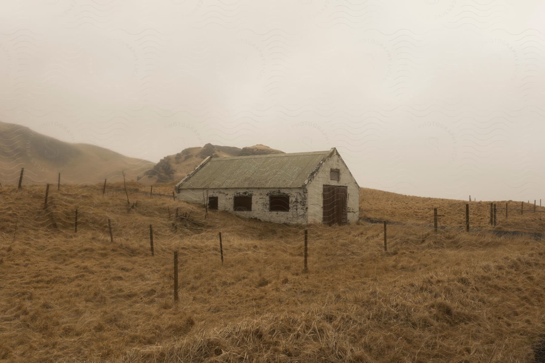 An old barn on a hillside covered in brown dead grass surrounded by fence posts
