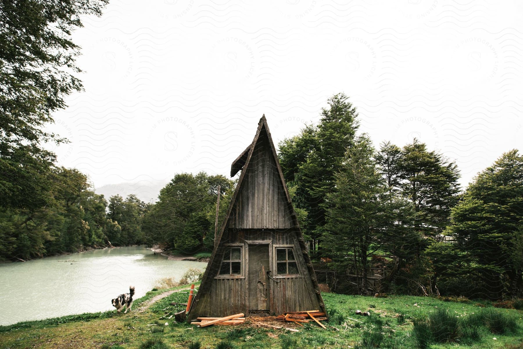 A small worndown triangular house sits beside a river in a field surrounded by trees on a cloudy summer day