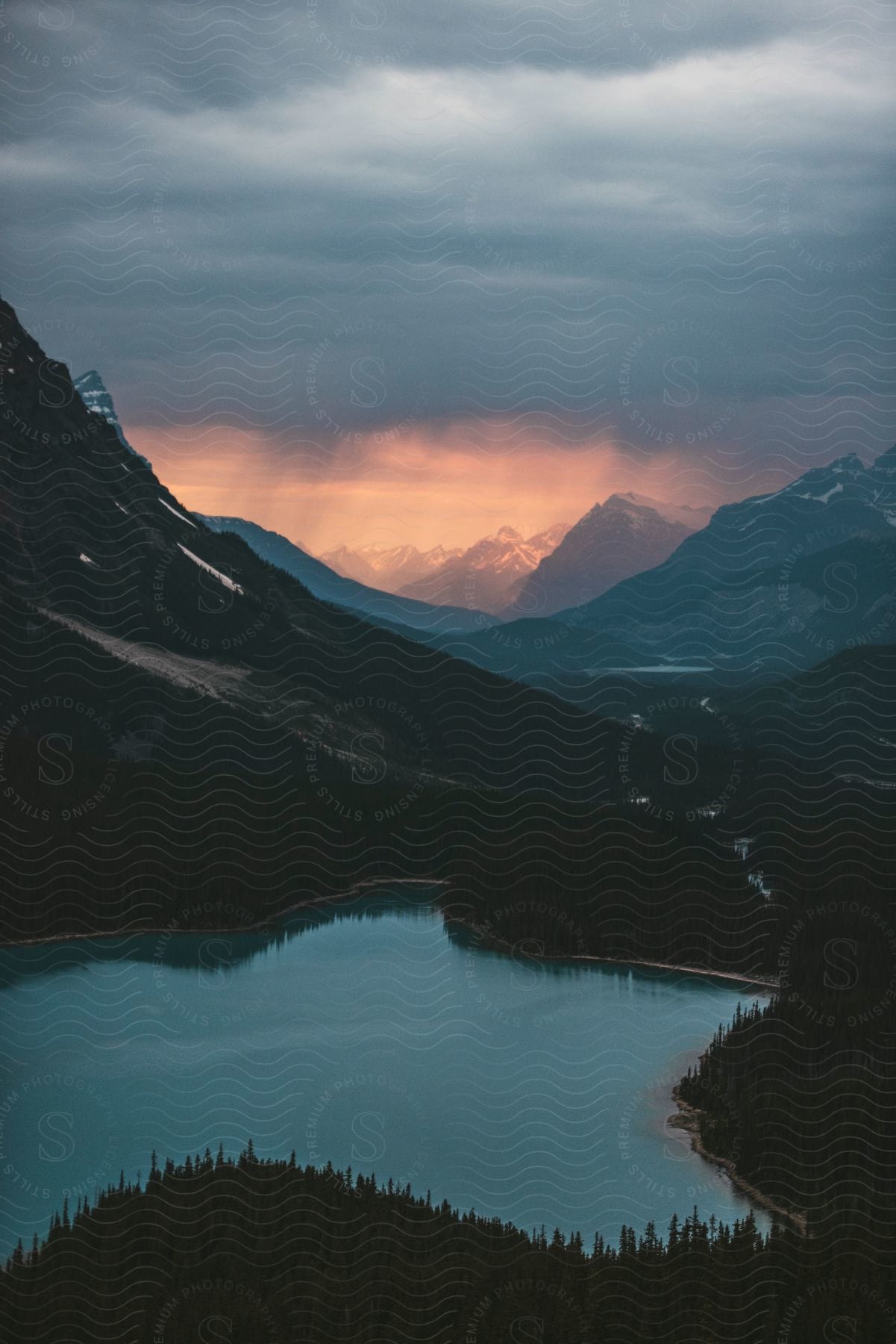 A forest of trees surrounds a mountain lake with mountains in the distance under a cloudy sunset sky