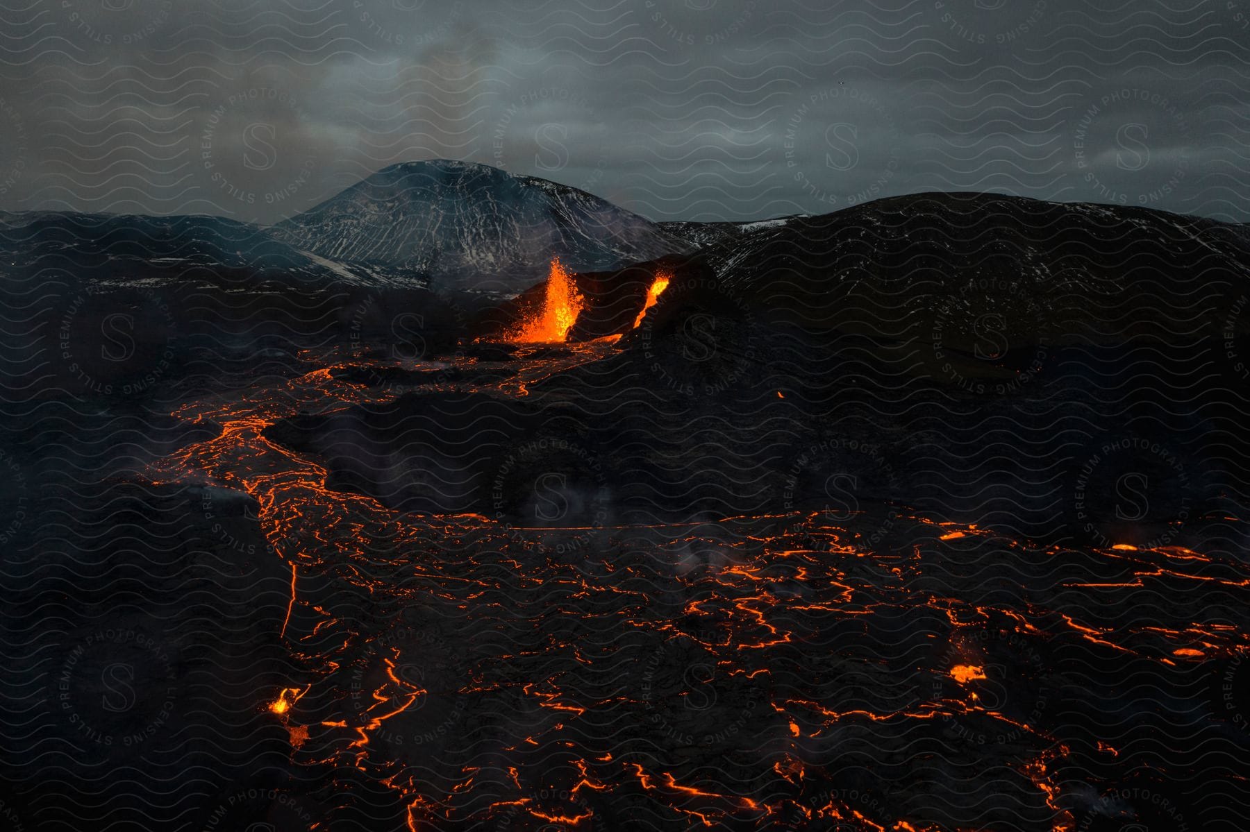 Lava bursts and flows from a volcano creating a fiery river under a cloudy night sky