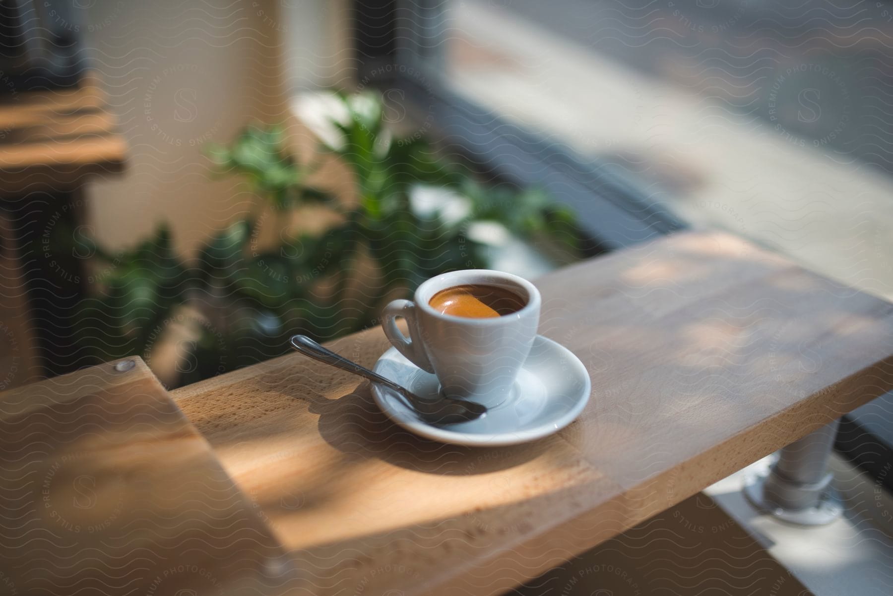 A cup of coffee with a spoon on the saucer sits on a counter near a window