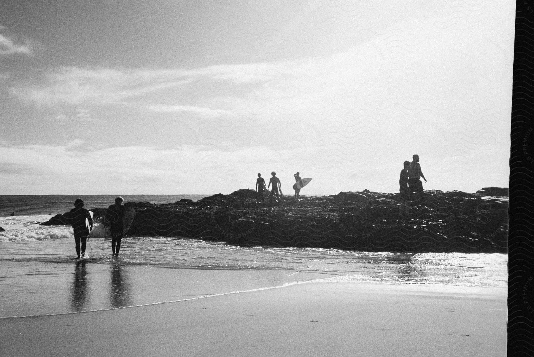 A group of people with surfboards walking on a sandy beach some on rocky terrain as ocean waves crash against the shore on a sunny day