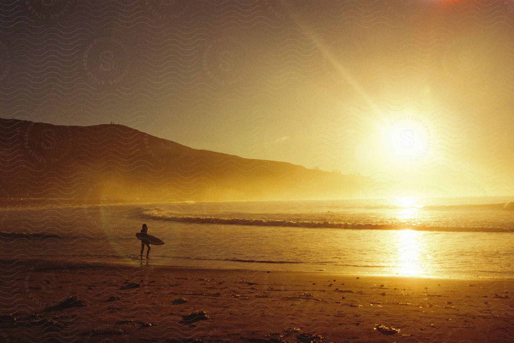 A person walks along the shoreline at dusk with a mountain in the background and a surfboard in hand