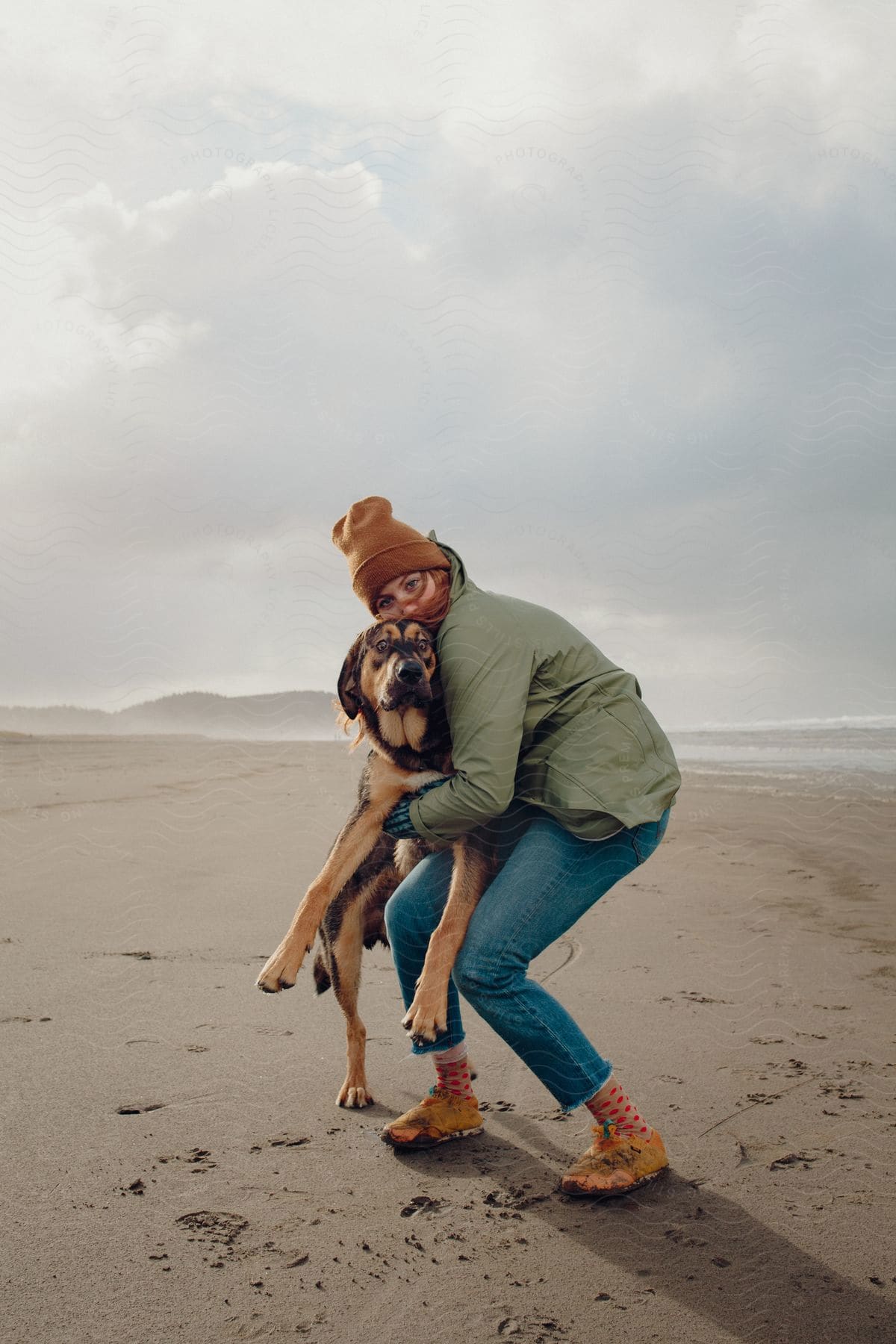 A woman with red hair wearing winter clothes hugs a dog at the beach