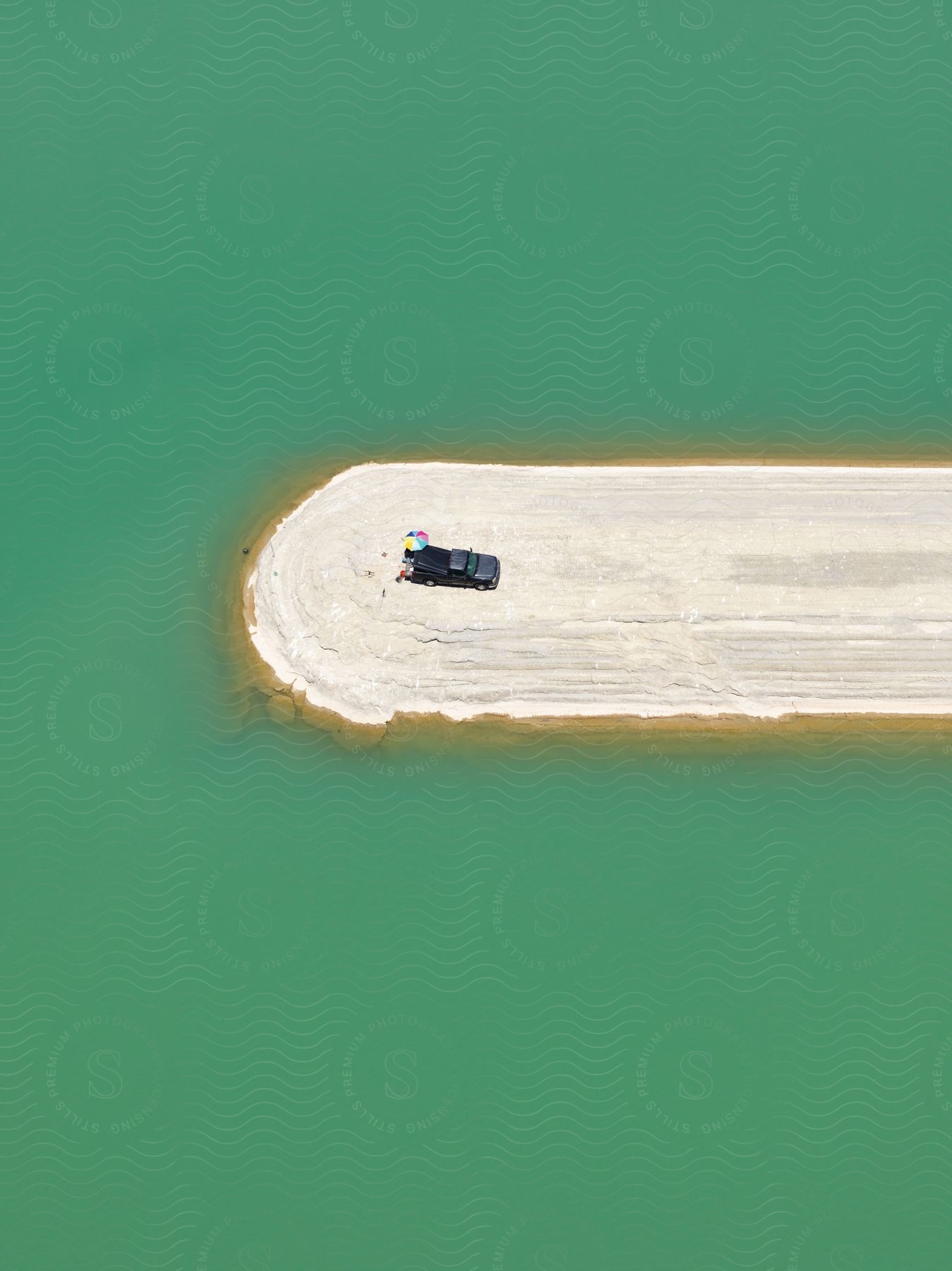 A truck is parked on a narrow sandy beach above the ocean with a white sandbar and multicolored beach umbrella