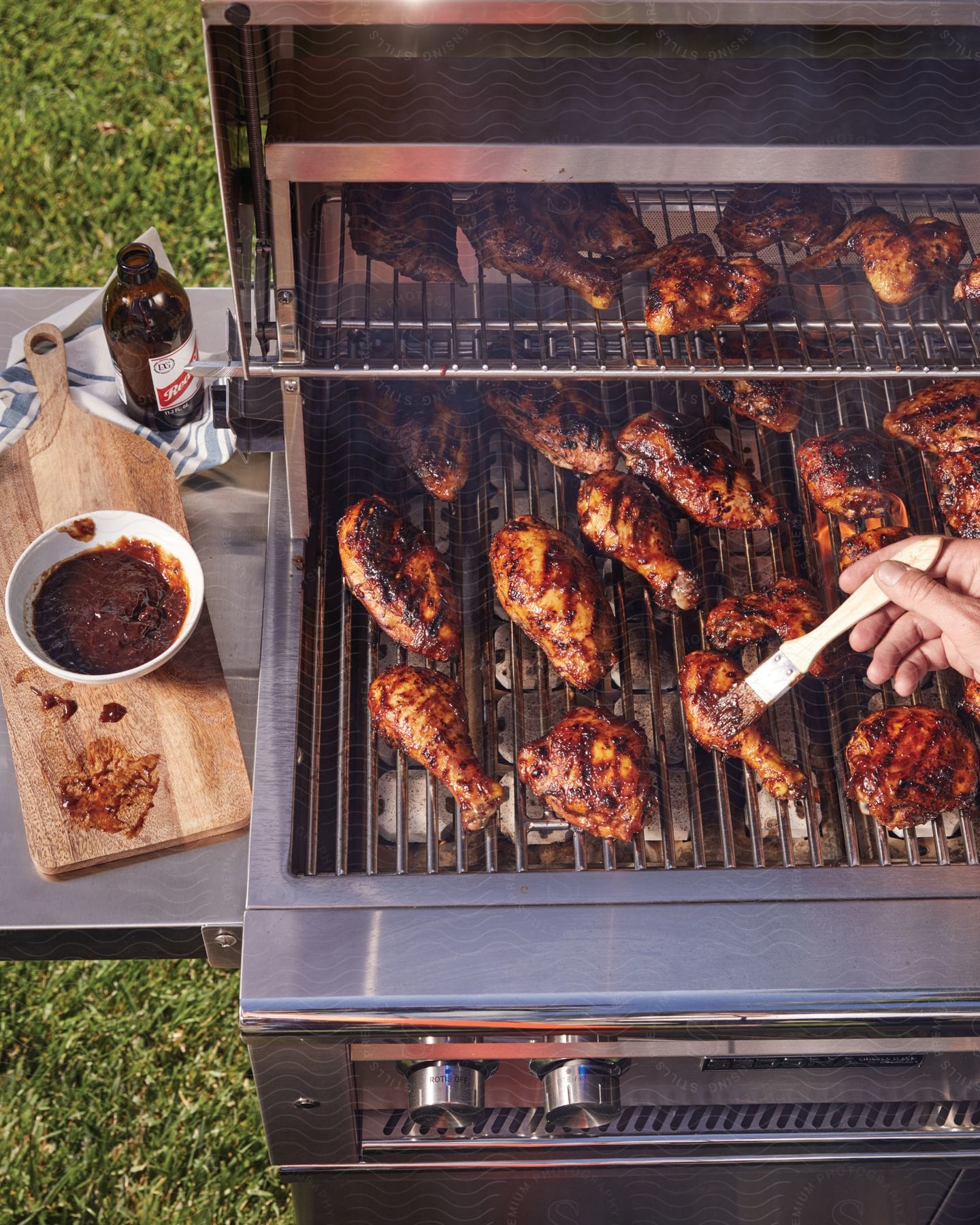 A person is using a hand to brush bbq sauce over chicken legs on a grill outdoors