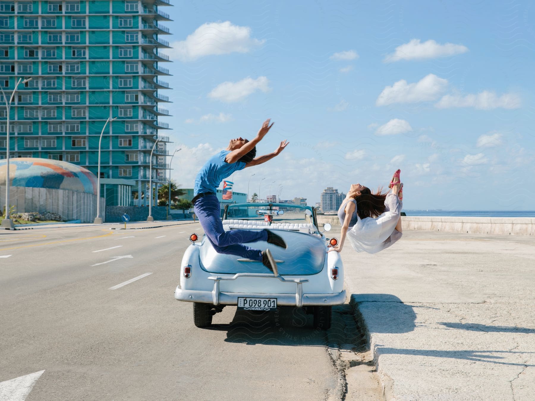 A couple jumps near a vintage car on a beachfront city