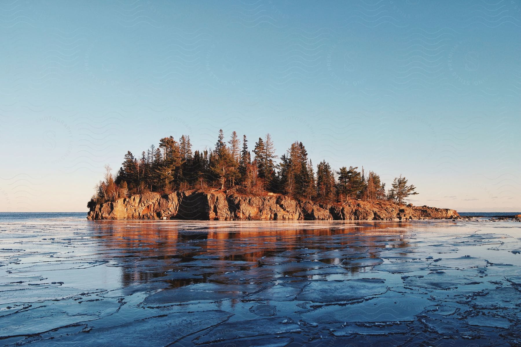 A rock island with brown trees in the middle of a cracked ice covered lake at daybreak