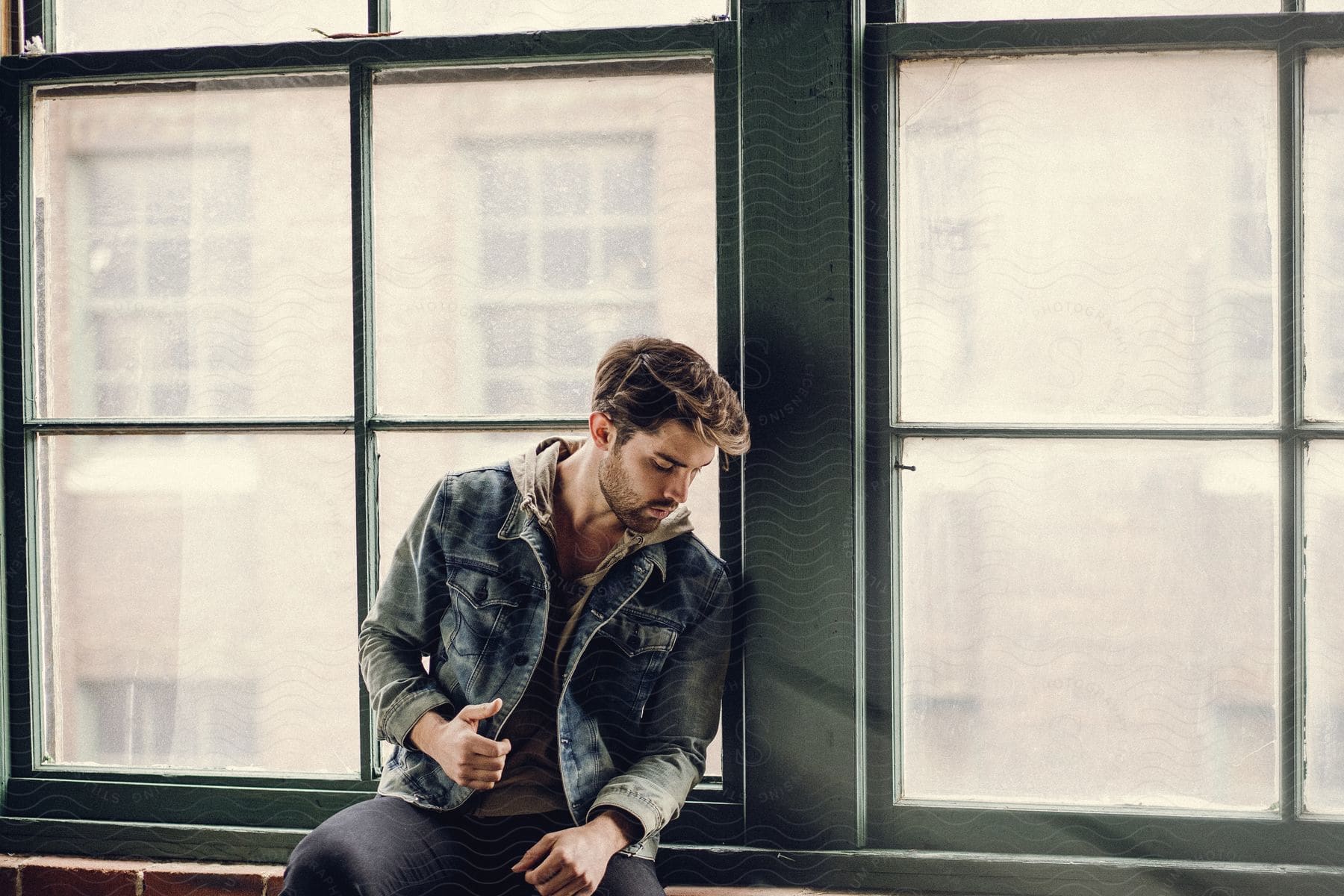 A man wearing a denim jacket sitting in front of a window posing for a photo