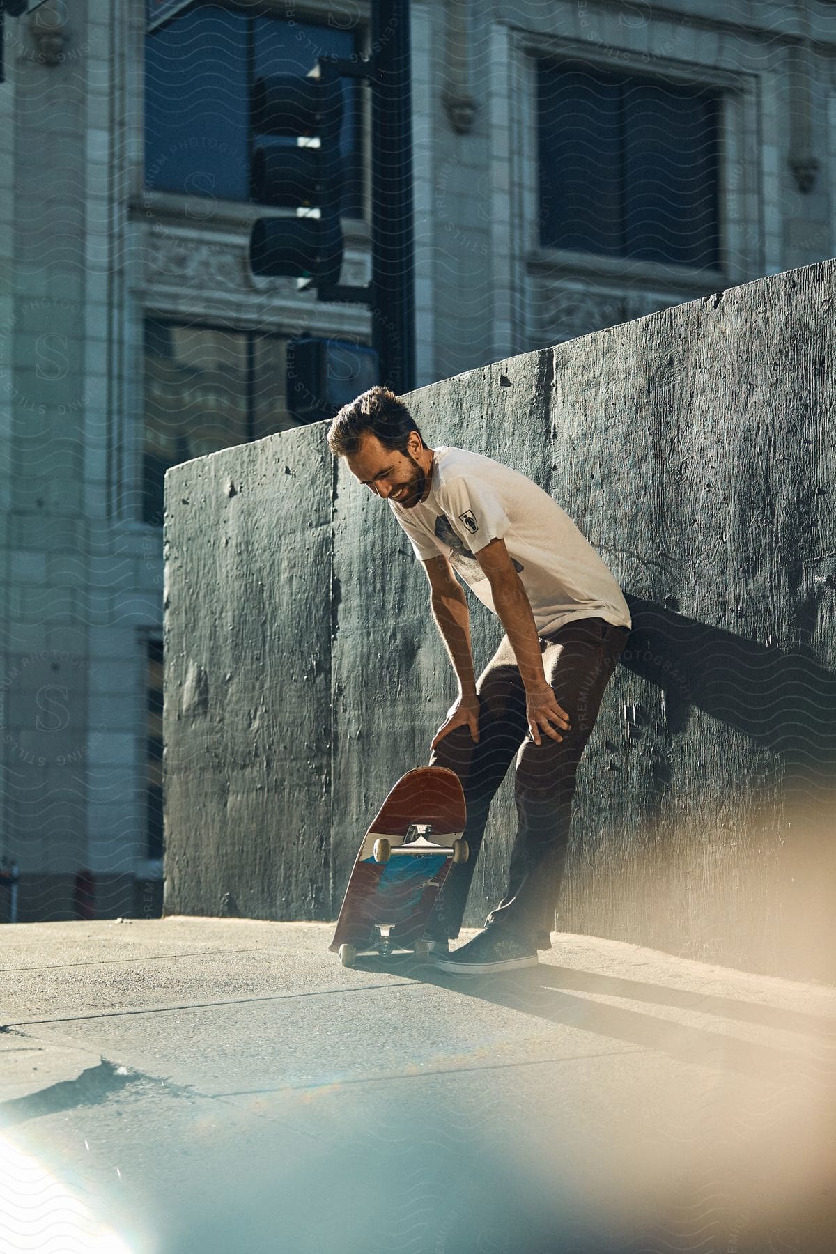 A man with a skateboard leaning against a wall with his hands on his knees