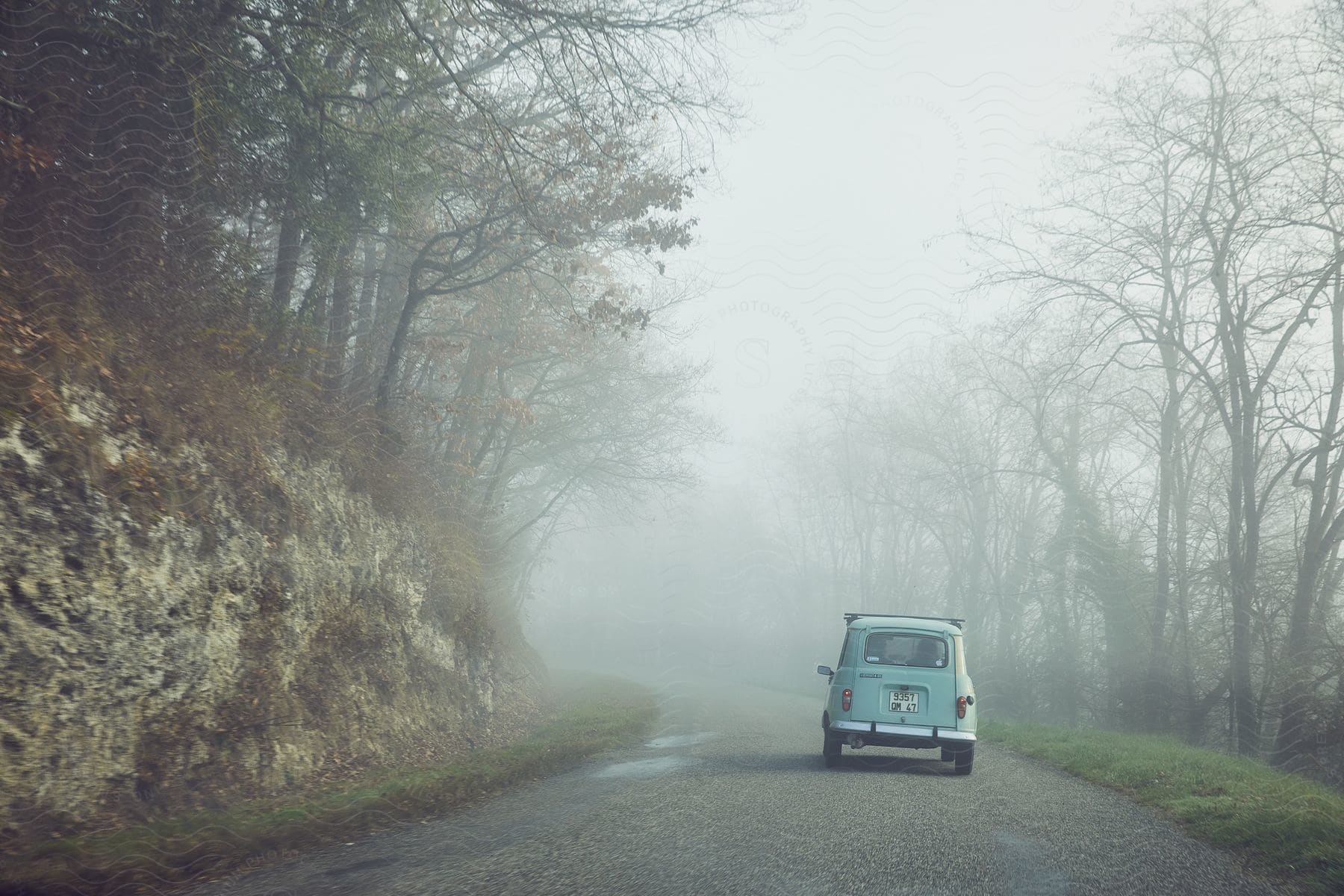 A small blue car is seen on a road surrounded by trees and fog