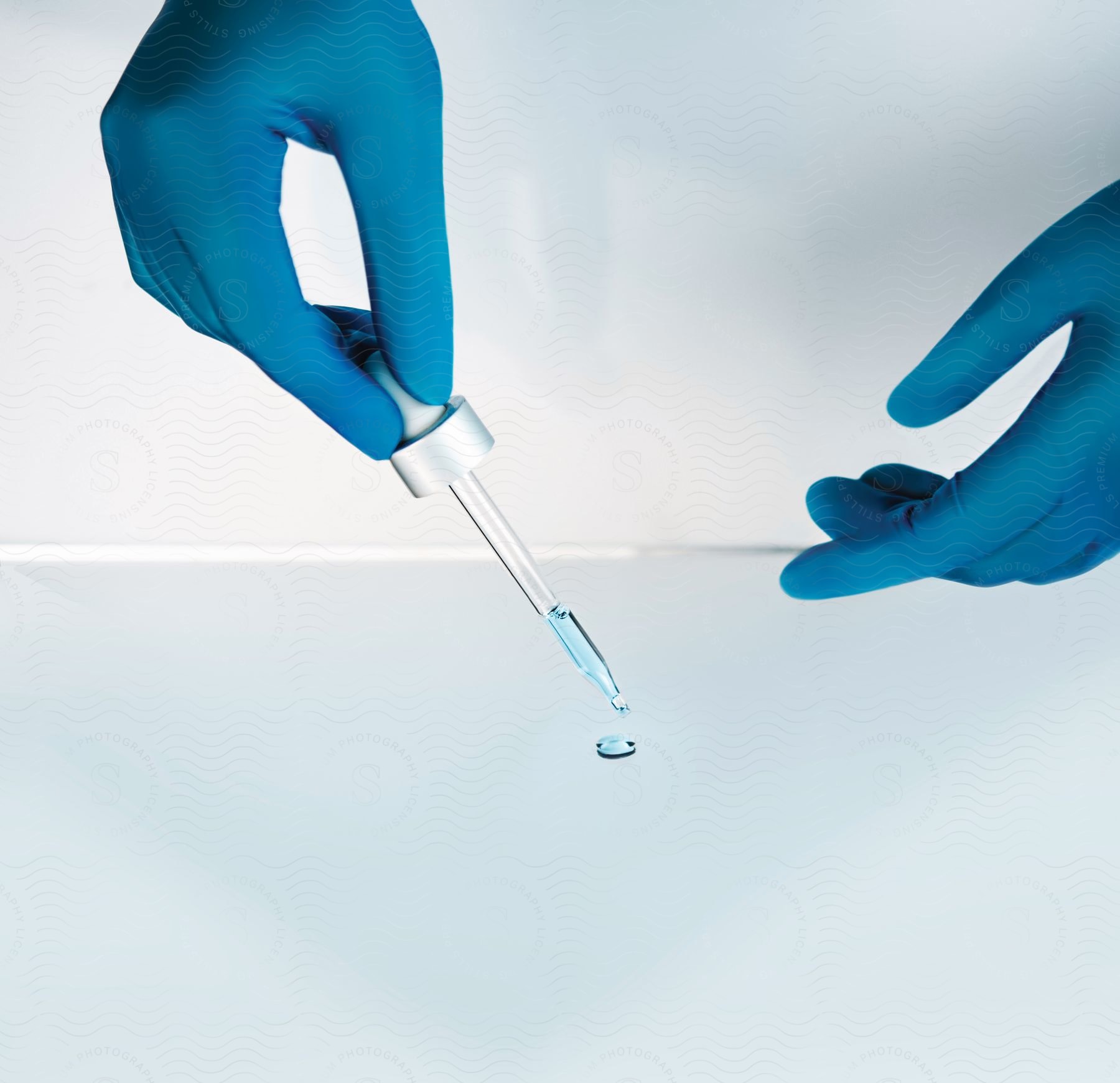 A pair of hands wearing blue gloves holds a dropper filled with clear blue liquid over a glass table