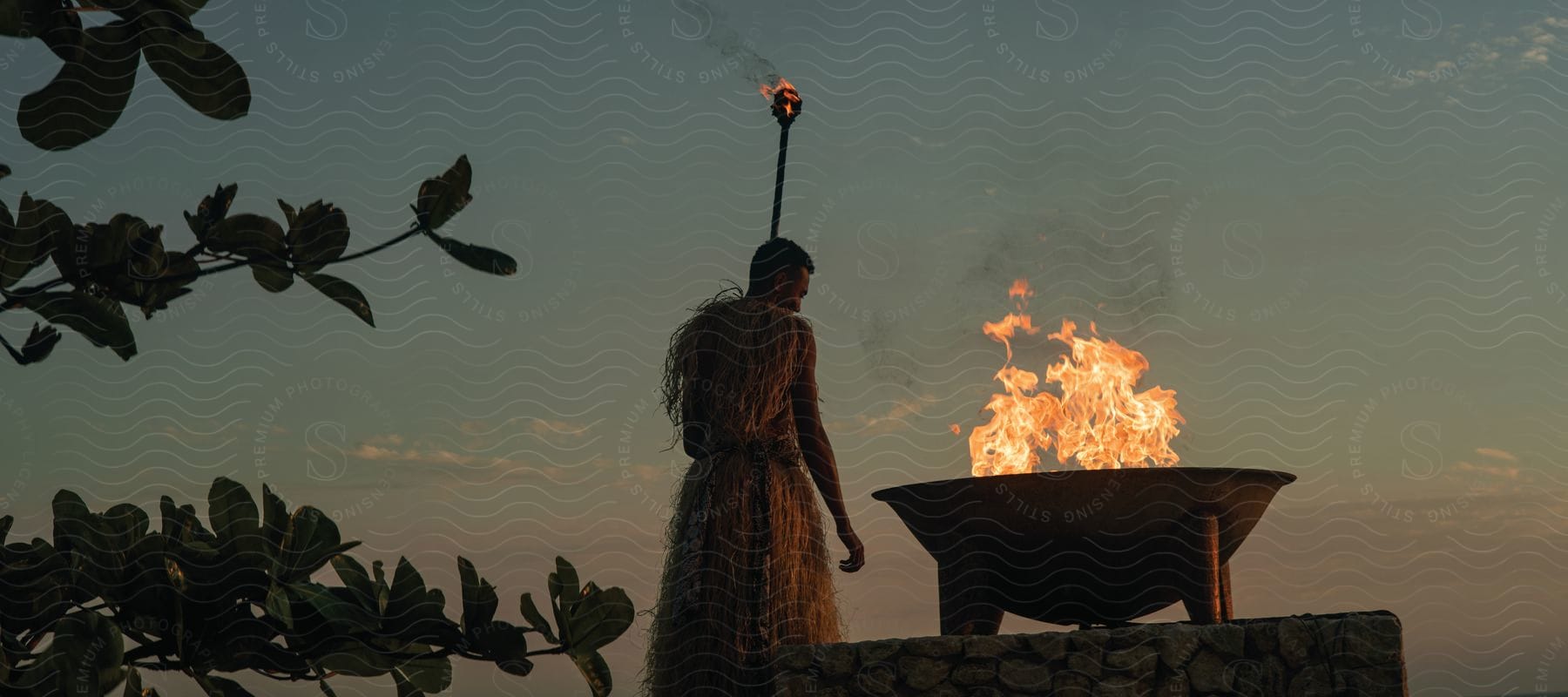 A man in costume holds a lit torch while standing outdoors near a fire