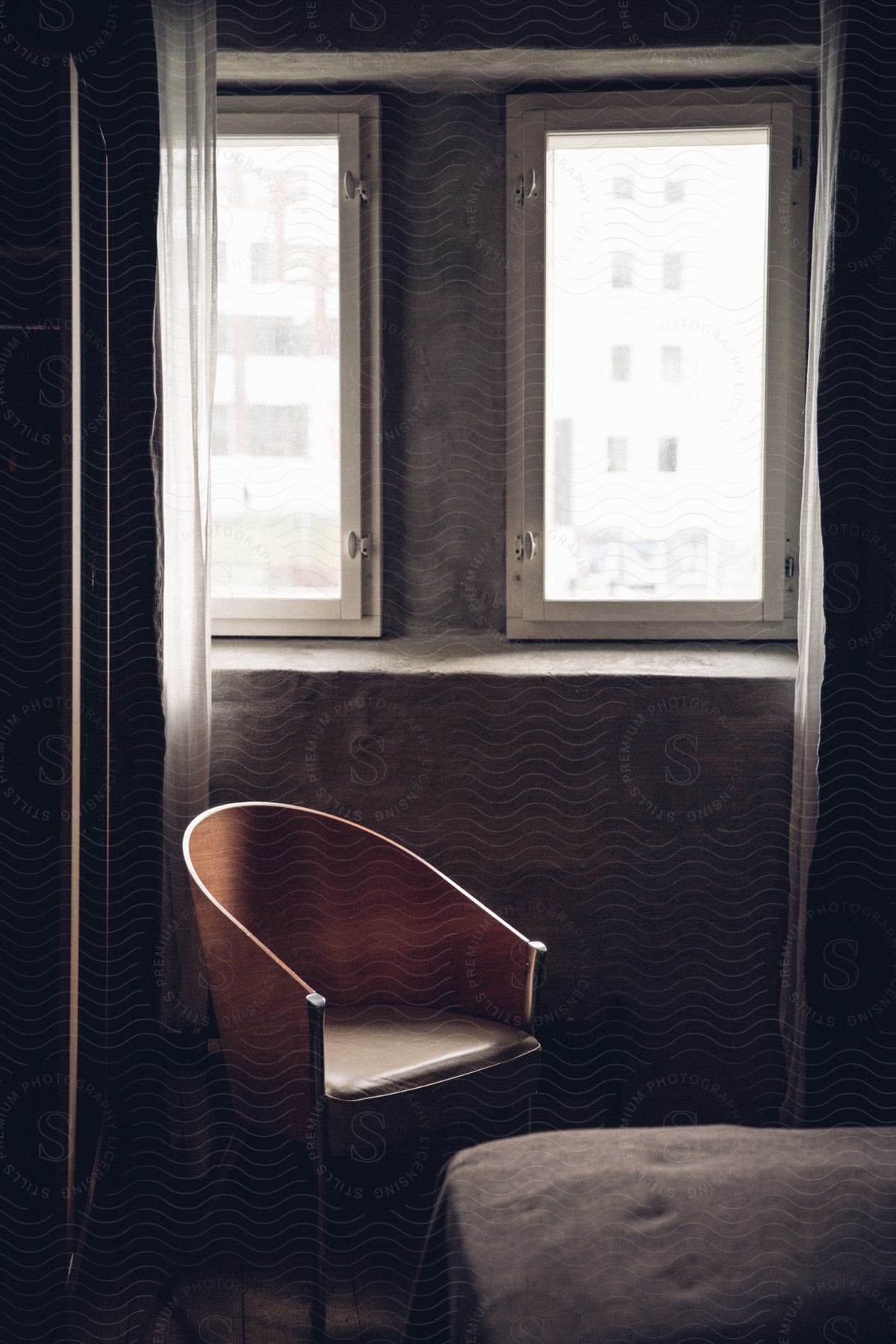 Interior of a small room with a bed a curved back chair and daylight coming through two windows with curtains