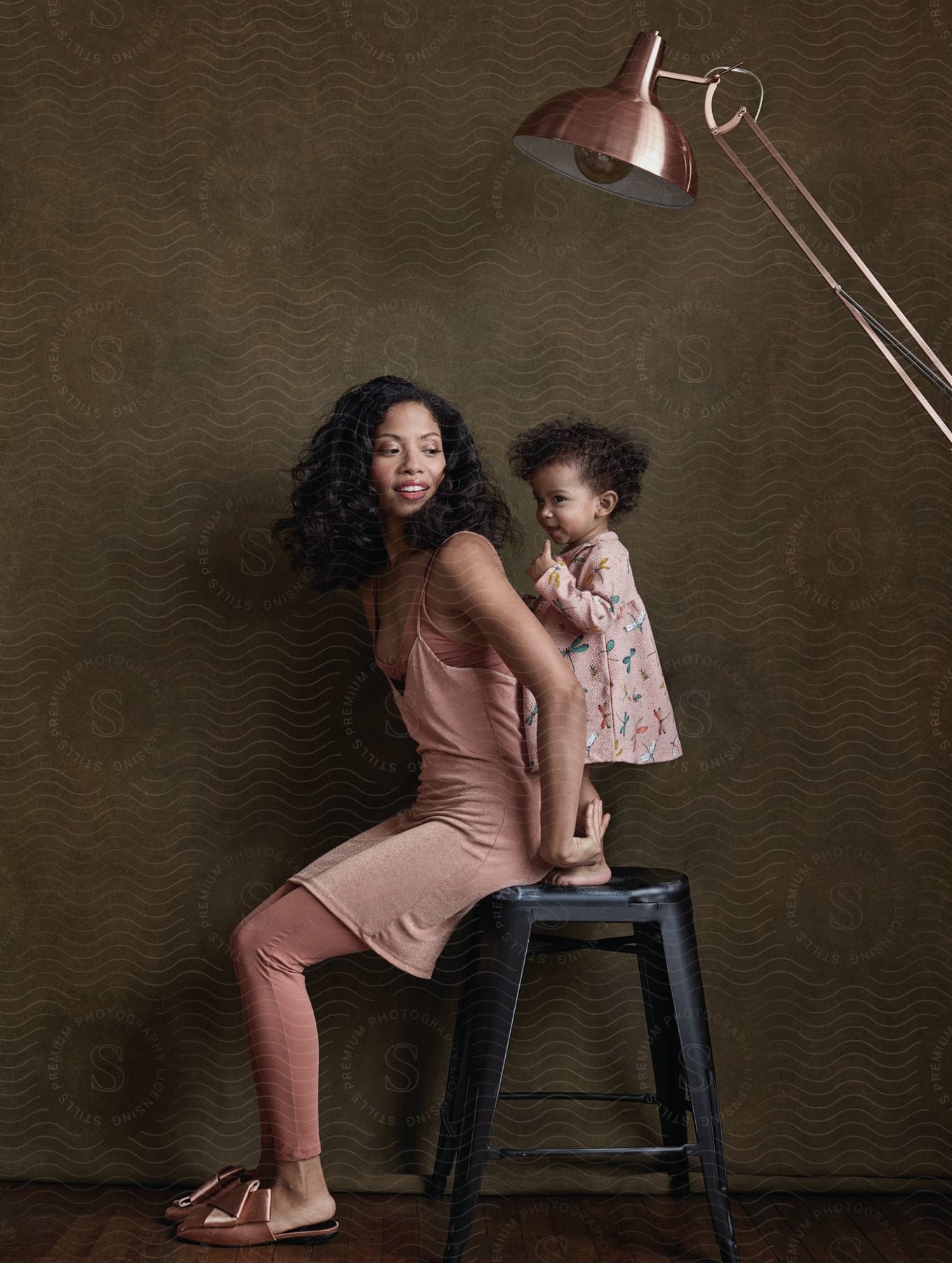 A mother and daughter dressed in pink are posed on a black stool under a pink lamp