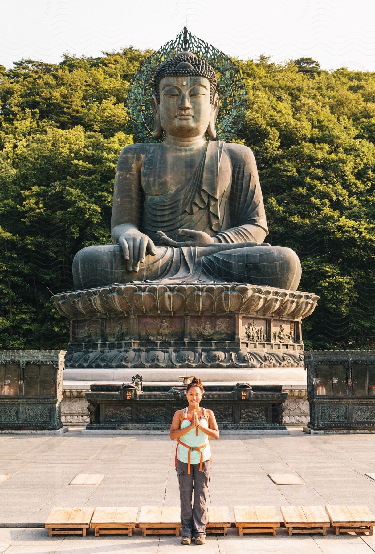 A woman in a green muscle shirt and pants stands in front of a statue of buddha in a park with a backpack slung over her shoulders her hands together in prayer looking towards the camera