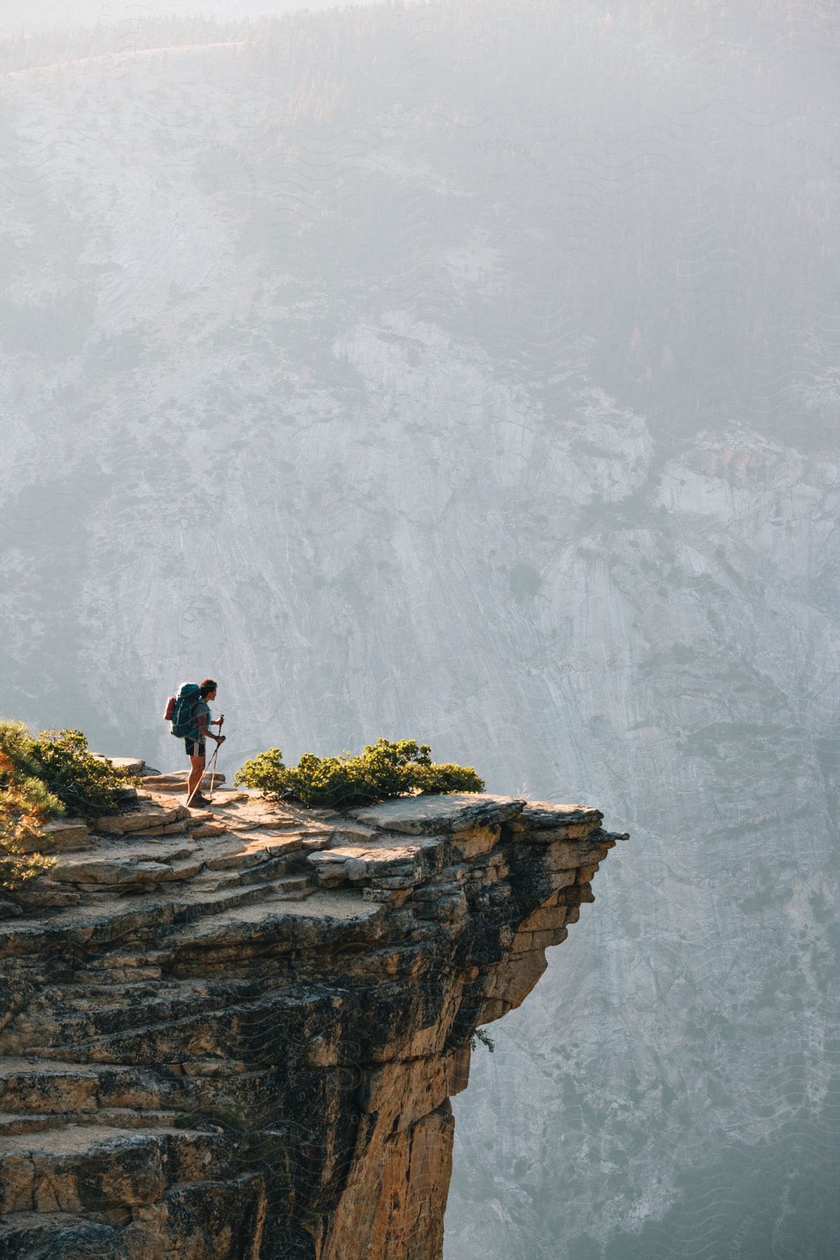 A hiker stands on a cliff edge gazing out at hazy mountains beyond holding hiking poles