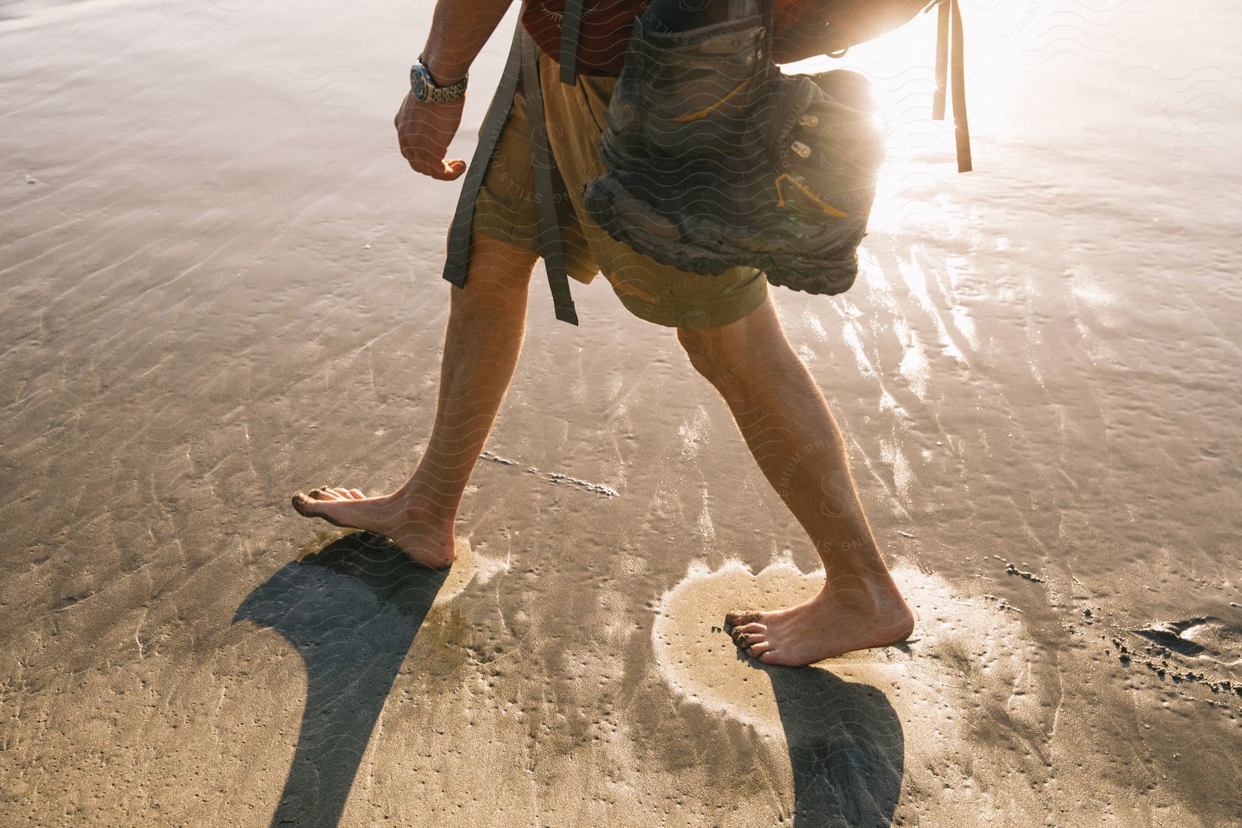 A man with hiking boots attached to his side walks barefoot on a beach at dusk or dawn